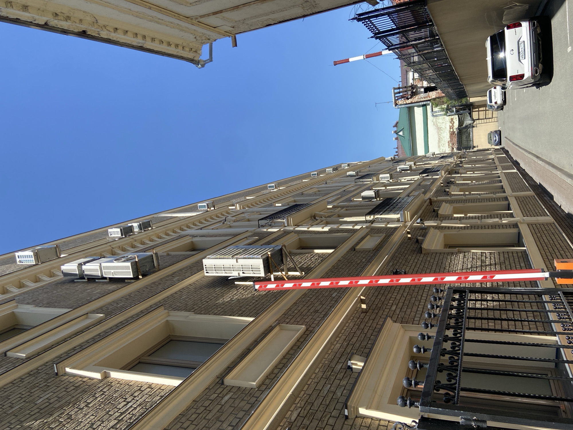 The image shows a view upwards within an urban alleyway in Kazan, Russia, characterized by buildings and infrastructure. A tall brick building with multiple windows, each topped with air conditioning units, forms one side of the alley. A red and white striped barrier is positioned near a wrought iron balcony. The sky is clear blue. On the opposite side of the alley, a parking area is visible, with parked cars. A distant smokestack with red and white stripes is also present. Architectural elements like cornices and ledges are visible on the tops of the buildings.