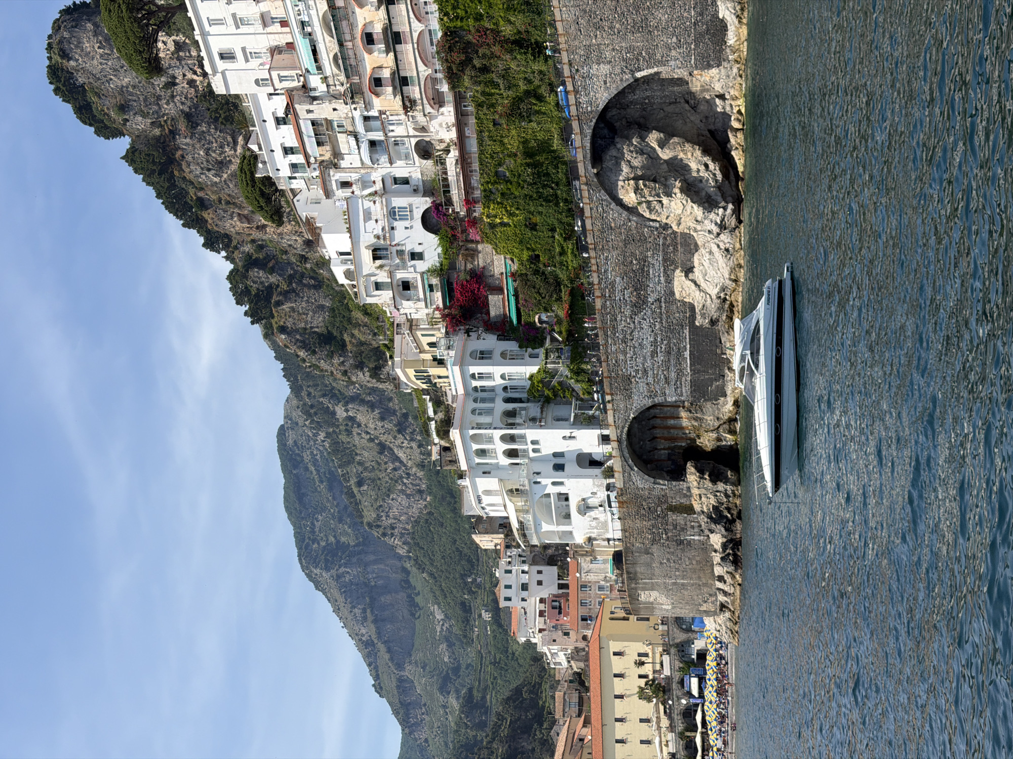 The image shows the coast of Pogerola, Italy. Tall, rugged cliffs flank the buildings, which are predominately white and stacked one above the other. In the foreground, a modern white boat is moored alongside a stone wall with arched openings. The sea extends to the right of the boat, reflecting the sunlight. Further below, a beach is visible, lined with rows of umbrellas. The sky is partially visible, displaying a light blue color.