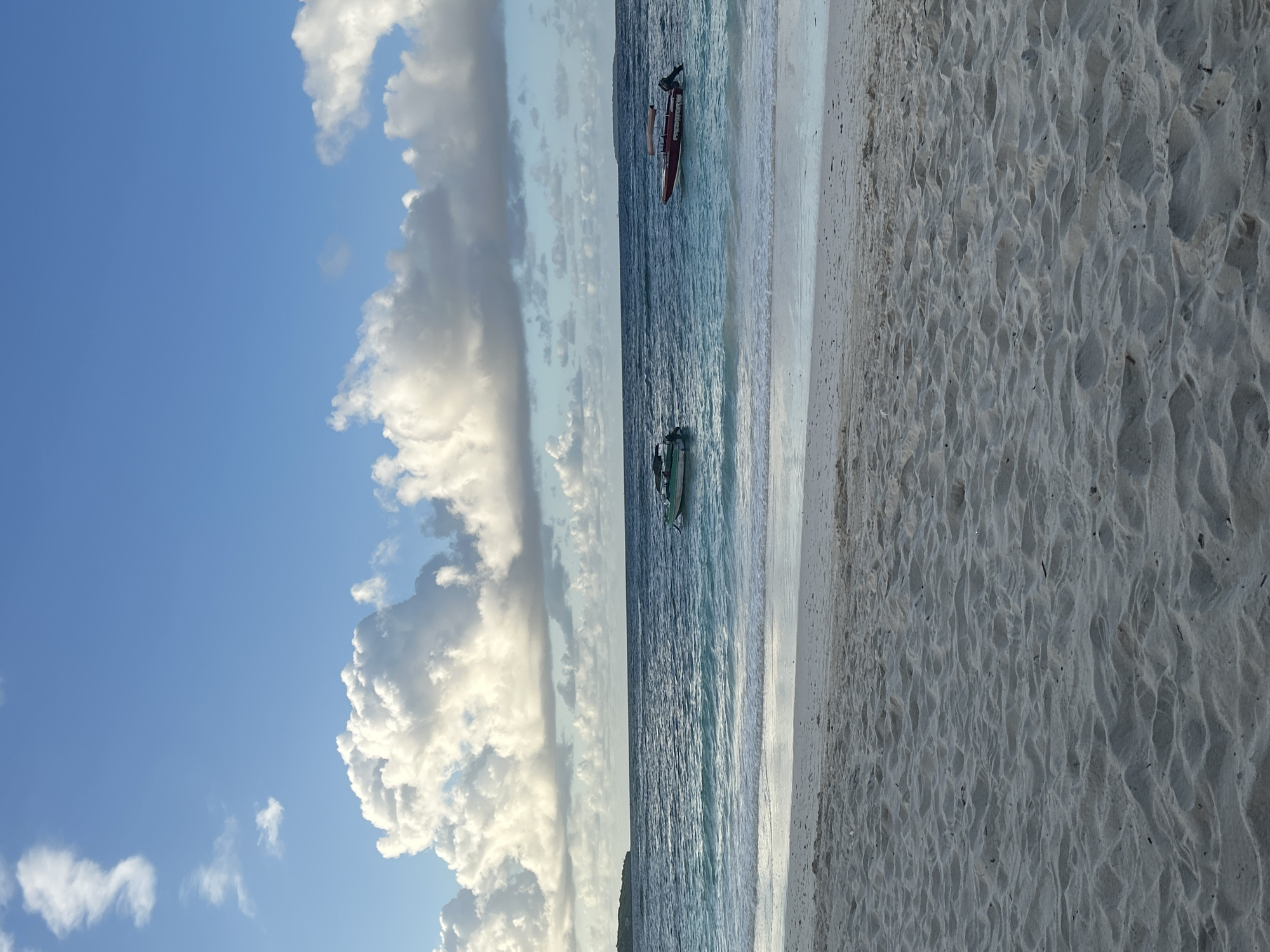 This image depicts a tranquil beach scene with two boats in the ocean. The foreground is dominated by the texture of white sand, showing ripples from the tide. The ocean, a vibrant turquoise and deep blue, stretches towards the horizon, with gentle waves lapping at the shore. Two boats are visible on the water: a smaller green boat is closer to the left side, while a larger red boat with a canopy is situated further to the right. In the distance, a faint landmass can be seen on the left, and the horizon is marked by the meeting of the sea and sky. The sky is a clear blue, dotted with fluffy white clouds, suggesting fair weather. The overall atmosphere is serene and picturesque. The image does not provide enough information to determine a specific location within the city of A R A, Indonesia, or to identify any people or specific actions.