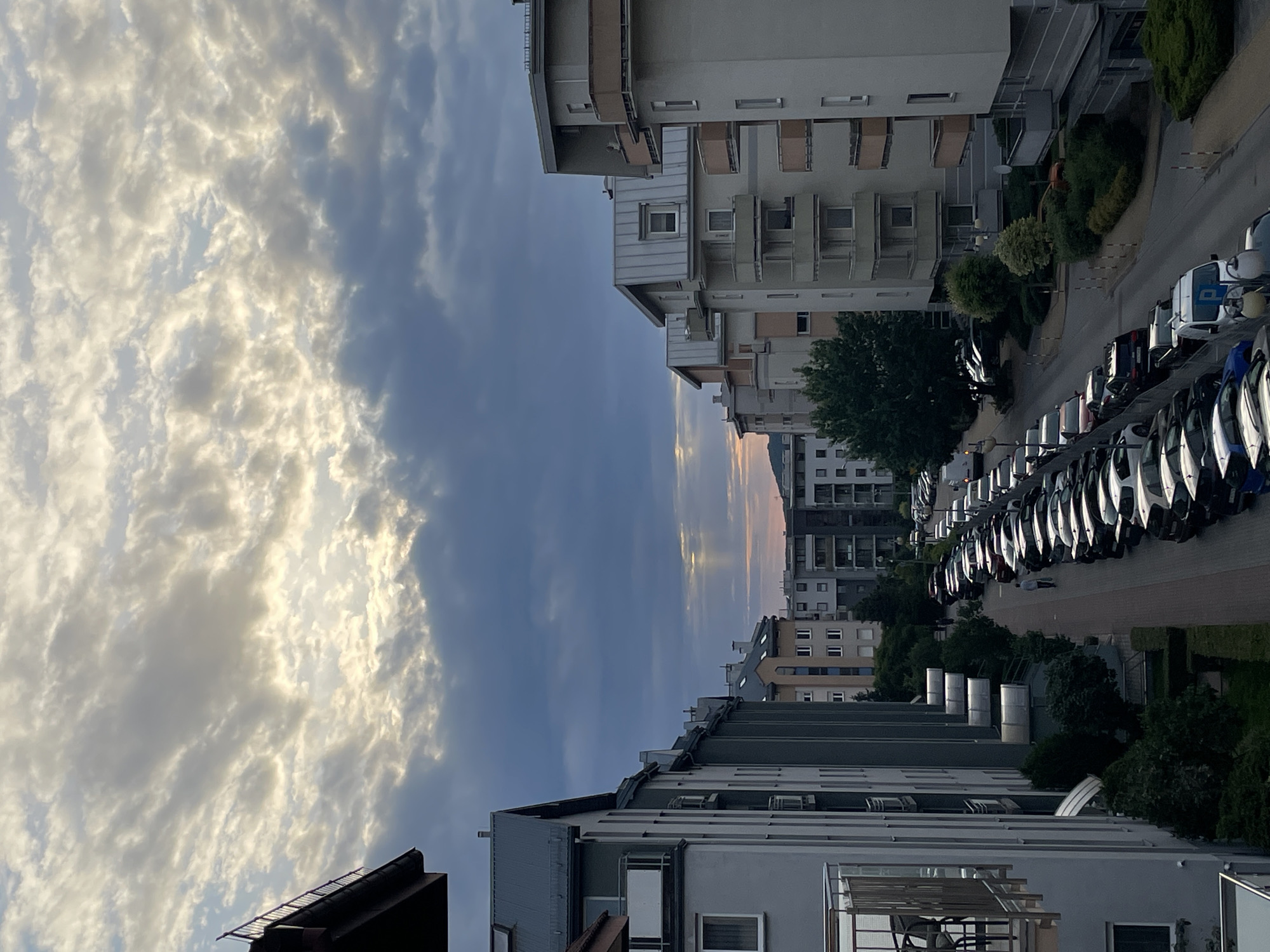 The image captures a street scene in an urban environment, likely at dusk, with apartment buildings lining both sides of the street. The sky is filled with dramatic clouds, with rays of sunlight breaking through in the distance. A long line of parked cars fills the street, with a few people visible walking along the sidewalk. The architecture of the buildings suggests a modern, possibly European, city. The overall impression is one of a typical residential street in a built-up area, with the natural beauty of the sky providing a striking contrast to the man-made structures.
