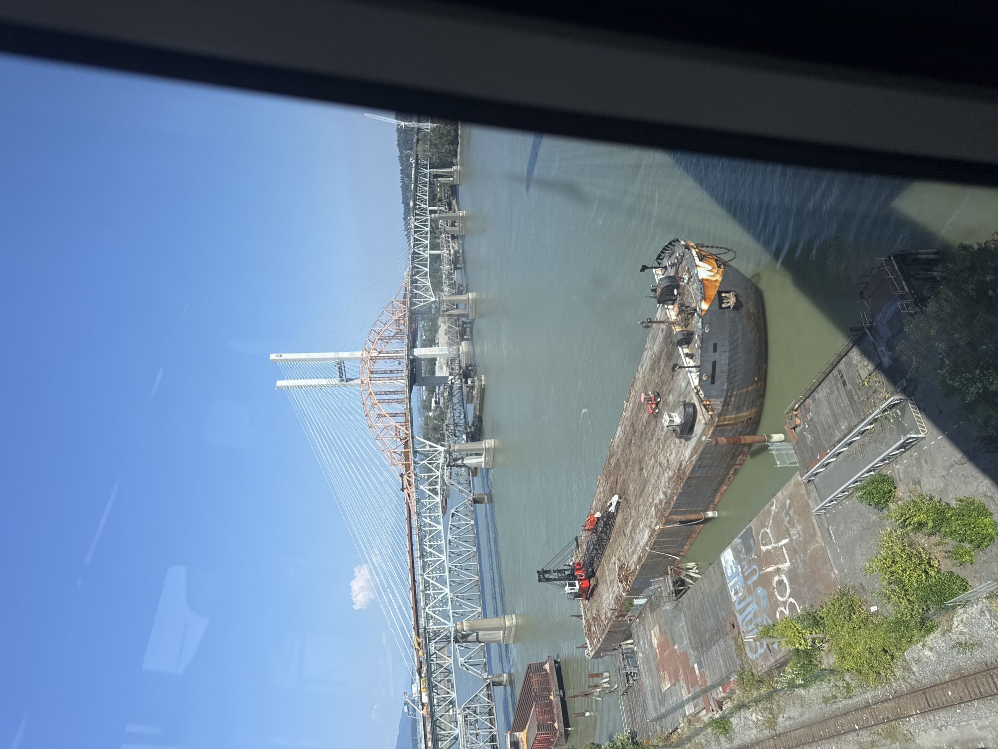An aerial, high-angle shot captures a wide, calm river in New Westminster, Canada, under a clear blue sky. In the foreground, a large, weathered barge with a crane on its deck is moored alongside a dock. The barge's surface is rusty and appears to be undergoing some form of work or is disused. Beside the barge, a smaller barge filled with what looks like cargo containers is also docked. Further in the background, multiple bridges span the river. The closest bridge is a substantial railway bridge with a lattice steel structure supported by concrete pillars. Beyond that, a modern cable-stayed bridge with white towers and orange and white cables dominates the skyline. The river is a muted green color, and the surrounding landscape features trees and a distant urban setting. The image is framed by a dark, vertical element on the right side, possibly part of a window or structure.