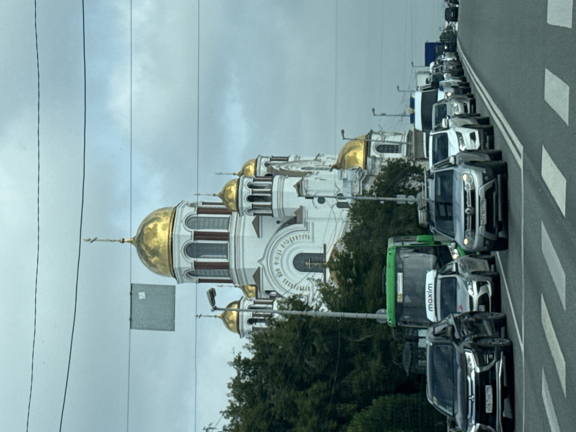 In Yekaterinburg, Russia, a queue of vehicles is stopped at a crosswalk. The vehicles include a black Mitsubishi Outlander, a white car with a "maxim" logo, and several other cars and a bus. In the background, a white church with golden domes, identified as the Church on the Blood in Yekaterinburg, stands prominently against a cloudy sky. The church is a historical landmark, built on the site of the Ipatiev House, where the last Russian Tsar Nicholas II and his family were executed. The scene suggests a typical urban traffic situation with the iconic church serving as a backdrop.