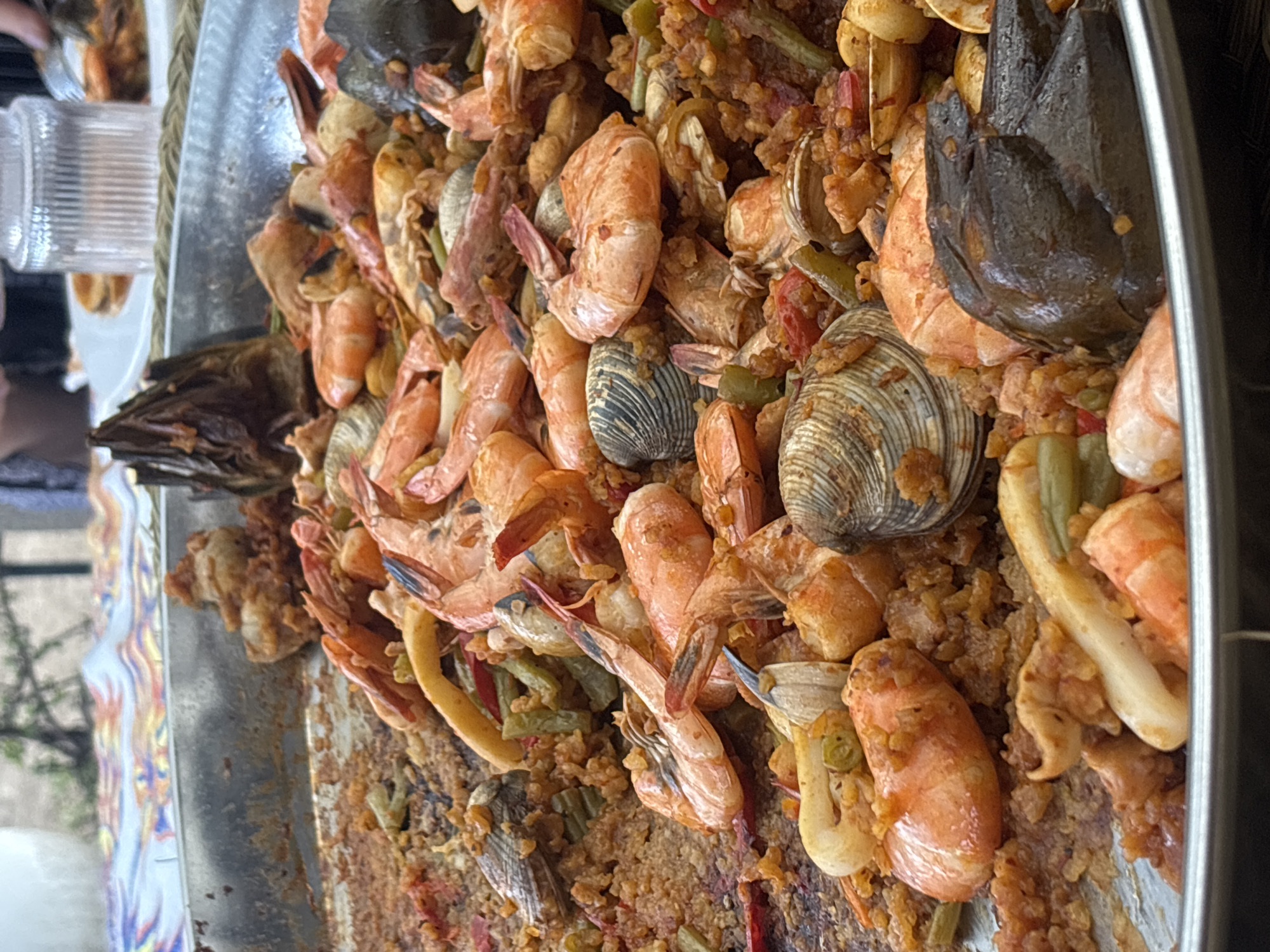 A close-up, angled shot depicts a large tray of paella. Prominent ingredients include numerous shrimp, clams, green beans, and what appears to be artichoke. The paella rests in a silver-colored tray with a patterned rim. A glass is also visible on the left of the frame. While the specific location within Puebla, Mexico is unknown, the dish itself suggests a culinary setting.