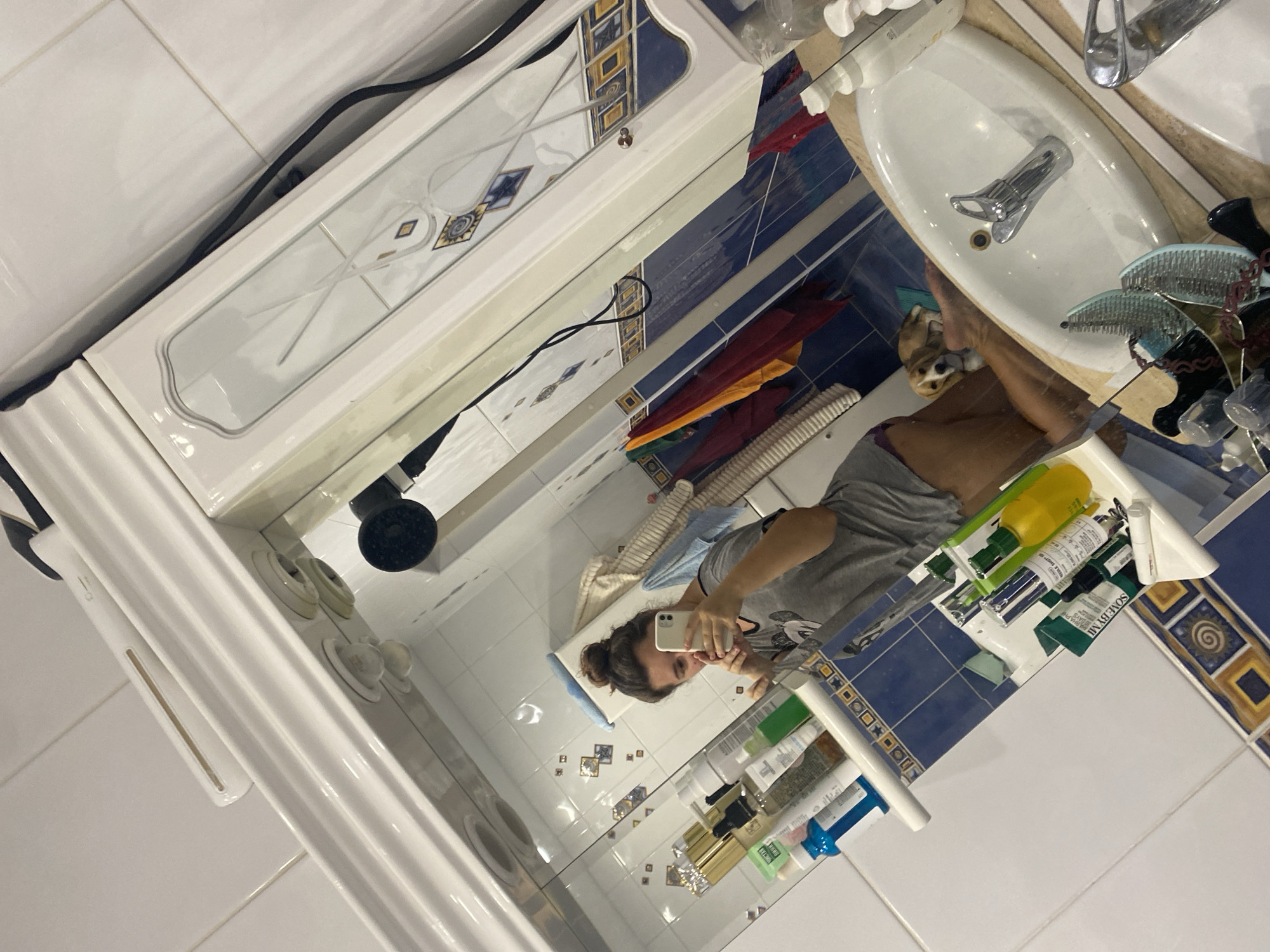 The image shows a person taking a selfie in a bathroom mirror. The bathroom appears to be well-lit and has tiled walls with blue and white patterns. A white cabinet with mirrored doors is above the sink. On the counter, various toiletries and bottles are arranged. A small dog is visible in the reflection, lying near the sink. The overall impression is that of a domestic bathroom setting. The image does not contain any specific information or visual cues that would indicate its location in the city of Perm, Russia, or any other specific geographic location.