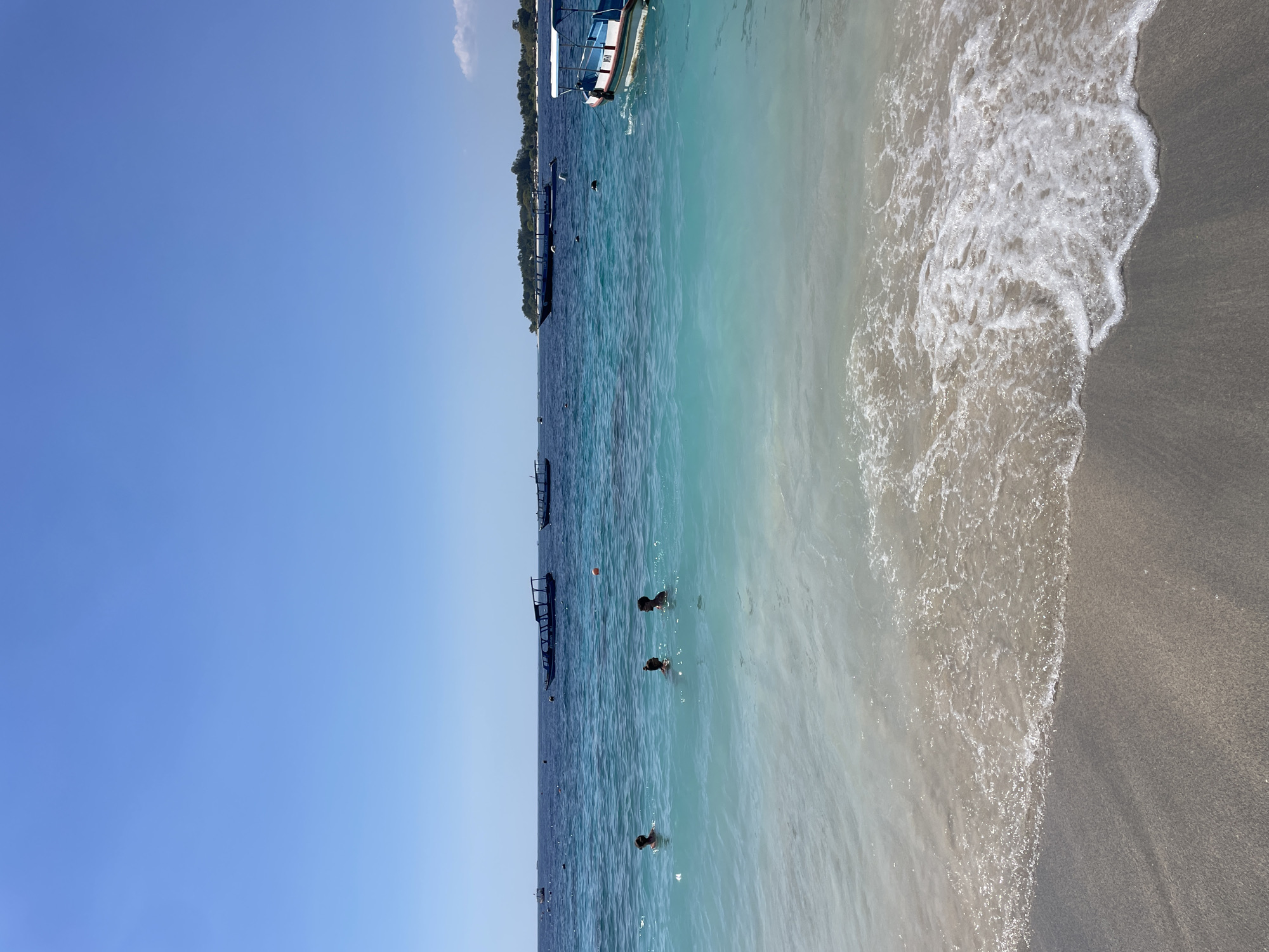 An idyllic view of a pristine beach in Indonesia unfolds under a clear, brilliant blue sky. The foreground is dominated by the gentle lapping of turquoise waves against a sandy shore, the water transitioning from a light, sandy hue near the beach to a deeper, mesmerizing blue further out. The water is clear, allowing visibility of the sandy bottom in the shallows. In the middle ground, three individuals are visible in the water, engaged in swimming or wading. Their forms are partially submerged, suggesting a leisurely swim. Further in the distance, several traditional Indonesian boats, known as *perahu*, are anchored or cruising on the calm sea. These boats, with their distinctive outriggers and canopies, dot the horizon, adding to the quintessential Indonesian coastal scene. The overall impression is one of tranquility and natural beauty, a perfect day at the beach in a tropical paradise.