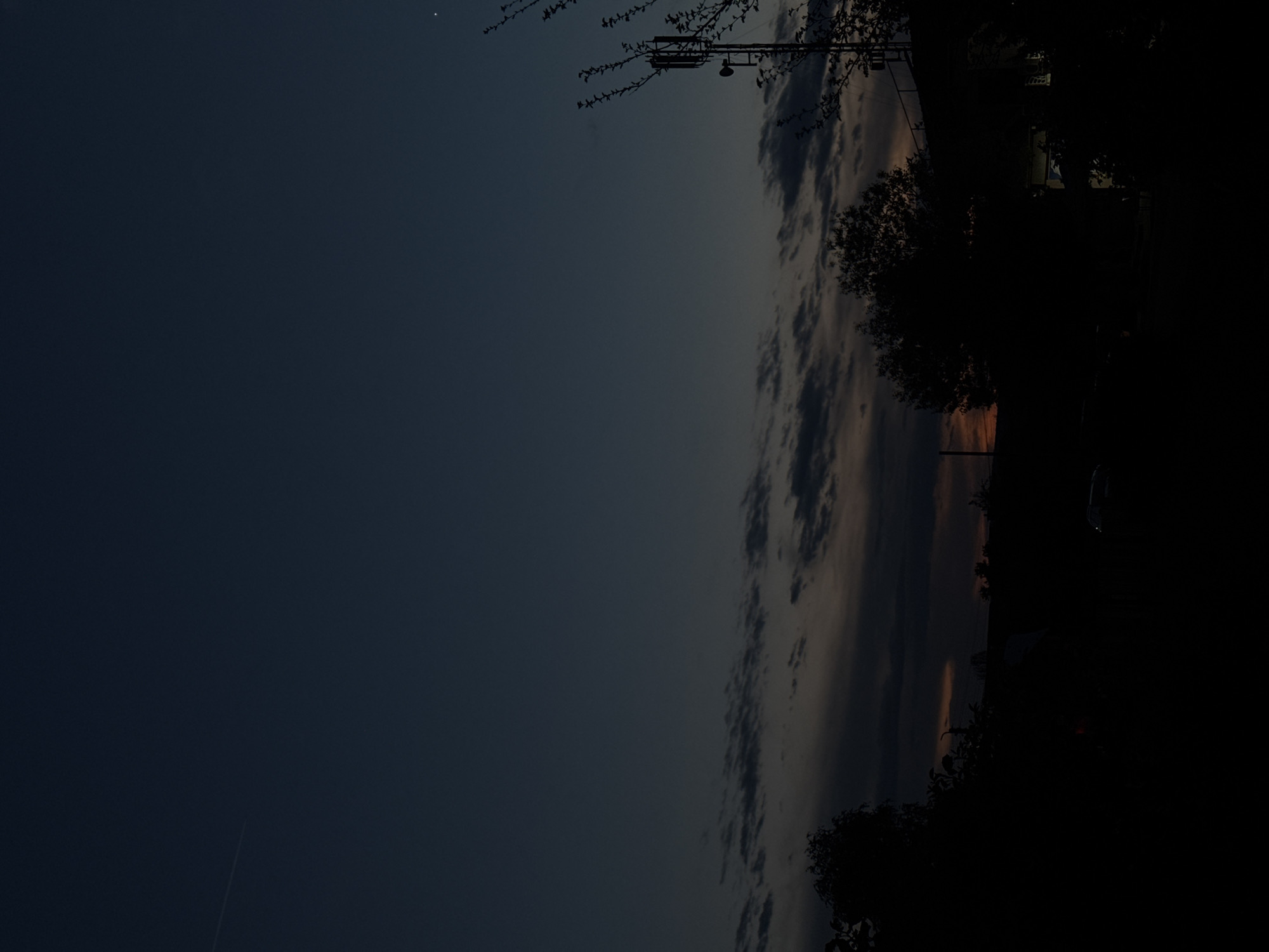 The image depicts a vertical view of the sky transitioning from a dark blue to a lighter hue near the horizon, possibly indicating dawn or dusk. Wispy clouds are visible along the horizon line. In the upper portion of the image, there is a silhouette of a utility pole or structure with branches of a tree surrounding it. The bottom and right side of the image are heavily shadowed, suggesting the presence of buildings, trees, or other tall structures that are blocking the light. A faint streak is visible in the dark blue sky.
The location in the city of Cherven Bryag, Bulgaria, cannot be determined based on the visual information provided in the image.