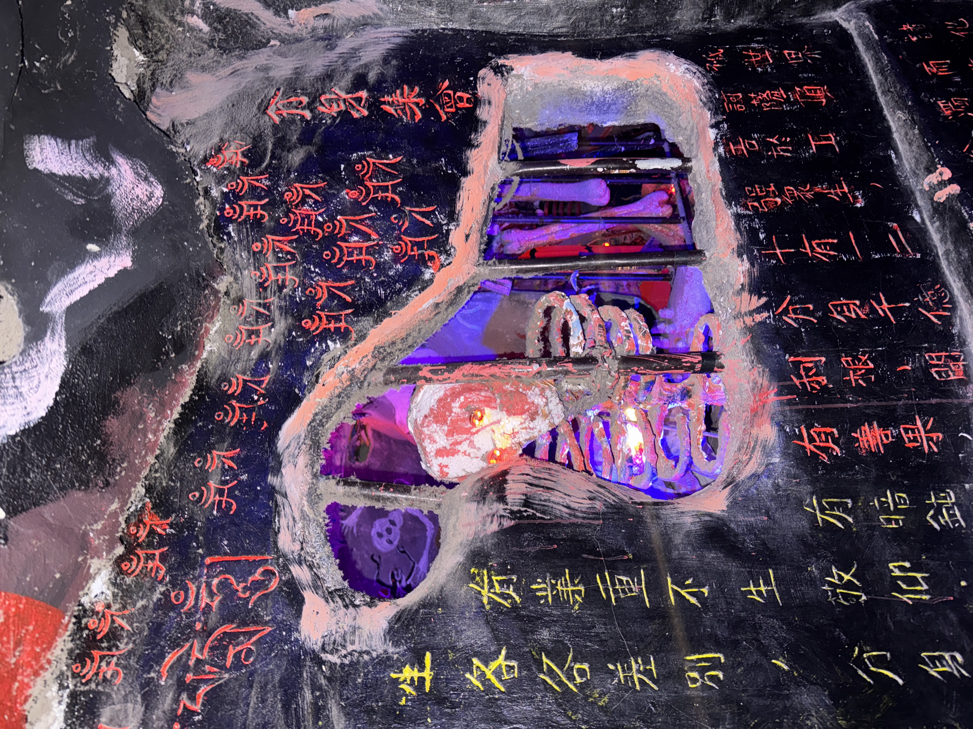 This media file captures a close-up of a multi-textured art installation, likely a wall-mounted piece, in Xuân Thành, Vietnam. The dominant feature is a dark, possibly black, surface resembling stone or heavily painted wood. Carved or built into this surface is a recessed area, roughly shaped like a stylized human torso, revealing internal components.
Inside the torso-shaped cavity, artistic representations of skeletal structures are visible. Metal bars form what appear to be ribs, and two skull-like objects are embedded within. One skull, painted reddish-white, is near the top of the cavity, while another, possibly darker with glowing eyes, is visible towards the bottom left. The interior is dramatically illuminated by purple and red artificial lights, casting an eerie glow on the skeletal elements.
The dark exterior surface is adorned with various inscriptions in both red and yellow characters. Some red characters are stylized, resembling ancient scripts like bone script or seal script, while others are more conventional Chinese or Vietnamese script. The yellow characters are primarily in a more standard block font. The visible text includes phrases such as "翁身集會" (Assembly of the Old/Elder Body), "有業重不生" (Heavy karma prevents rebirth), "眾生" (All living beings), "生各各差別,分身化" (Births each differ, transform into separate bodies/avatars), and "有善果, 有暗鈍" (There are good fruits, there is ignorance/dullness). These phrases suggest philosophical or spiritual themes, possibly related to Buddhist concepts of karma, rebirth, and the nature of existence.
The piece has a macabre yet contemplative aesthetic, blending elements of anatomy, spirituality, and artistic lighting. The setting appears to be indoors or in a controlled environment due to the artificial illumination, contributing to its enigmatic and profound atmosphere.