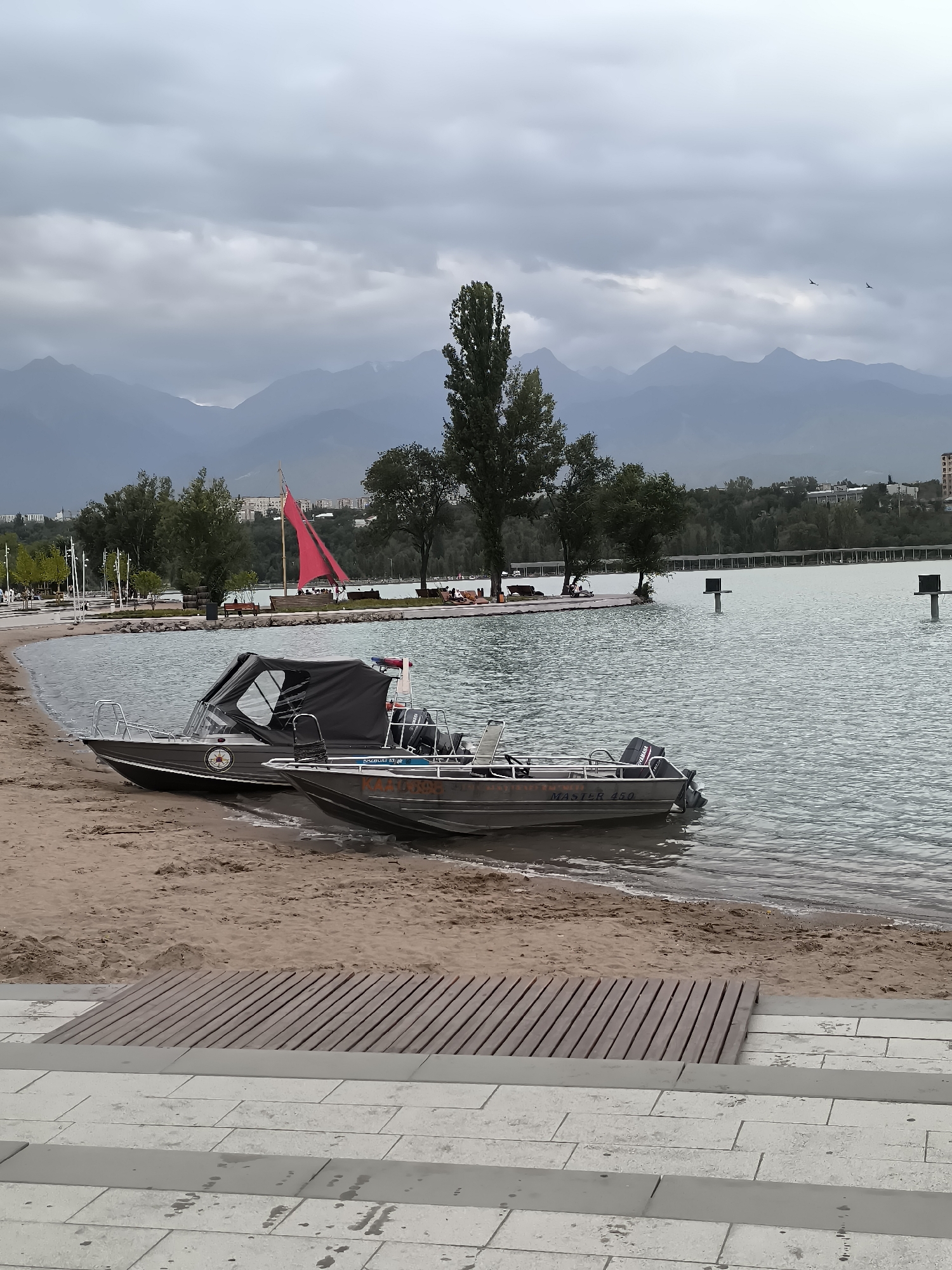 An overcast day at a man-made lake in Almaty, Kazakhstan, likely the Aqua Park or a similar recreational area. The foreground shows a concrete walkway with tiered steps and a wooden ramp leading to a sandy beach. Two motorboats are parked on the sandy shore, partially submerged in the water. The larger boat, with a dark canopy, has "MASTER 450" visible on its side. Beyond the beach, the lake stretches out with several poles protruding from the water, possibly for markers or lights. In the mid-ground, trees line the shore, and in the distance, a city skyline with buildings is visible against a backdrop of hazy mountains. A red sail is prominent on a small boat in the lake. Two birds are flying in the sky. The overall atmosphere is calm and somewhat subdued due to the cloudy weather.