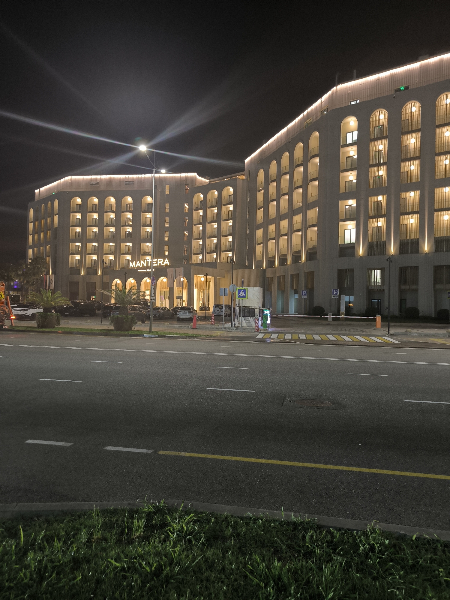 The image captures a nighttime view of the Mantera resort complex in Sirius, Russia. The resort is a large, multi-story building with a facade characterized by numerous arched windows, many of which are illuminated, suggesting occupancy. The building is lit from above with a warm-toned light. A street is visible in the foreground, with lane markings and a yellow line dividing traffic. On the right side of the image, a striped pedestrian crossing leads to the resort's entrance, where a barrier arm is lowered, indicating a controlled entry point, likely a parking or access control. Several cars are parked in front of the resort, and palm trees in planters add a touch of greenery. The sky is dark, with bright light streaks emanating from streetlights and possibly other sources, creating a somewhat dramatic nighttime atmosphere.
