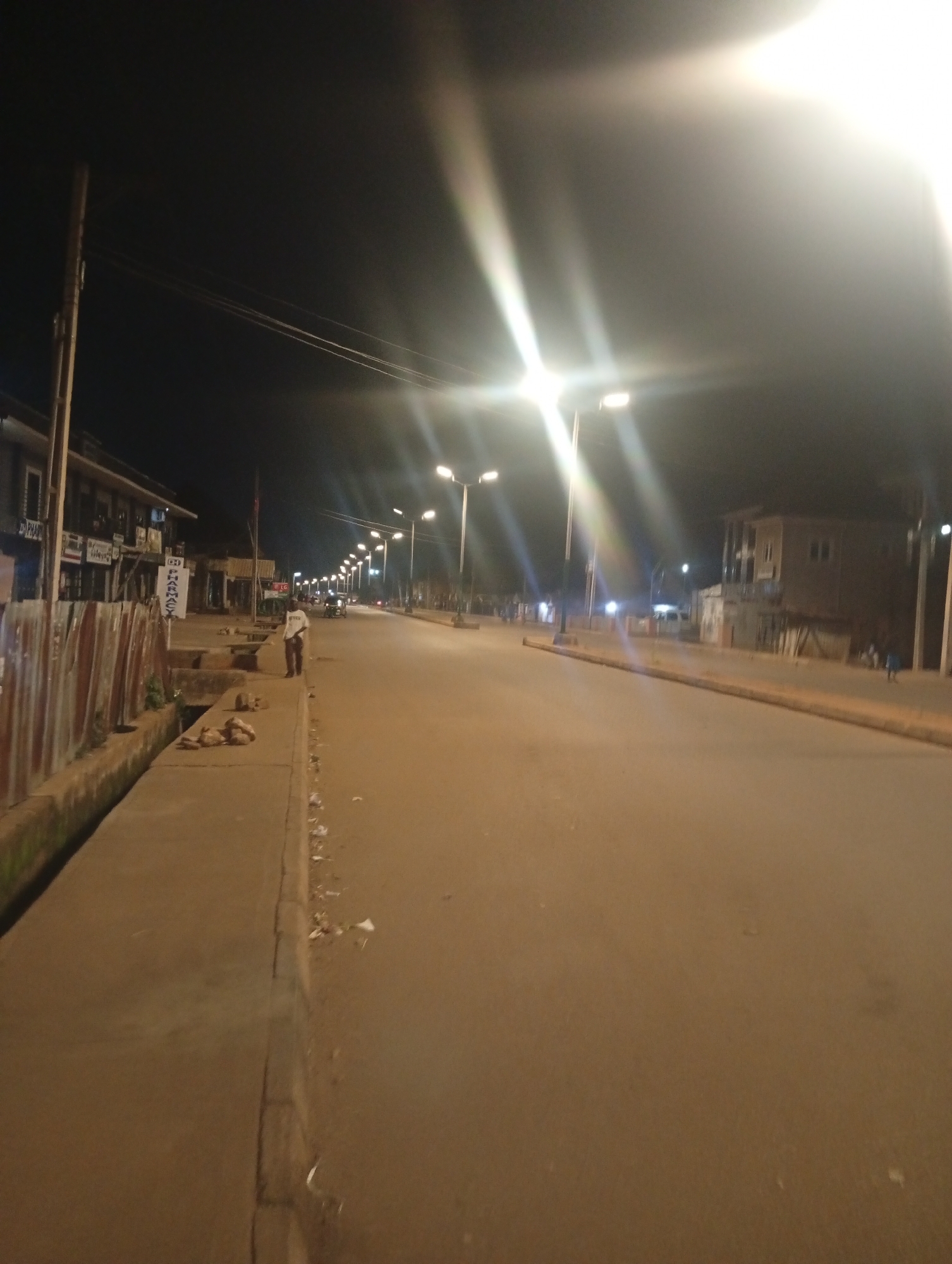 The image depicts a street scene in Bukuru, Nigeria, at night. The street is paved and appears to be a commercial area with buildings lining one side. Several streetlights illuminate the scene, creating long light streaks due to the long exposure. On the left side of the street, there is a sidewalk next to a drainage ditch. A person is standing on the sidewalk, looking towards the street. In the background, more streetlights stretch down the road, and other buildings and structures are visible. There is some debris scattered on the sidewalk and the road.
