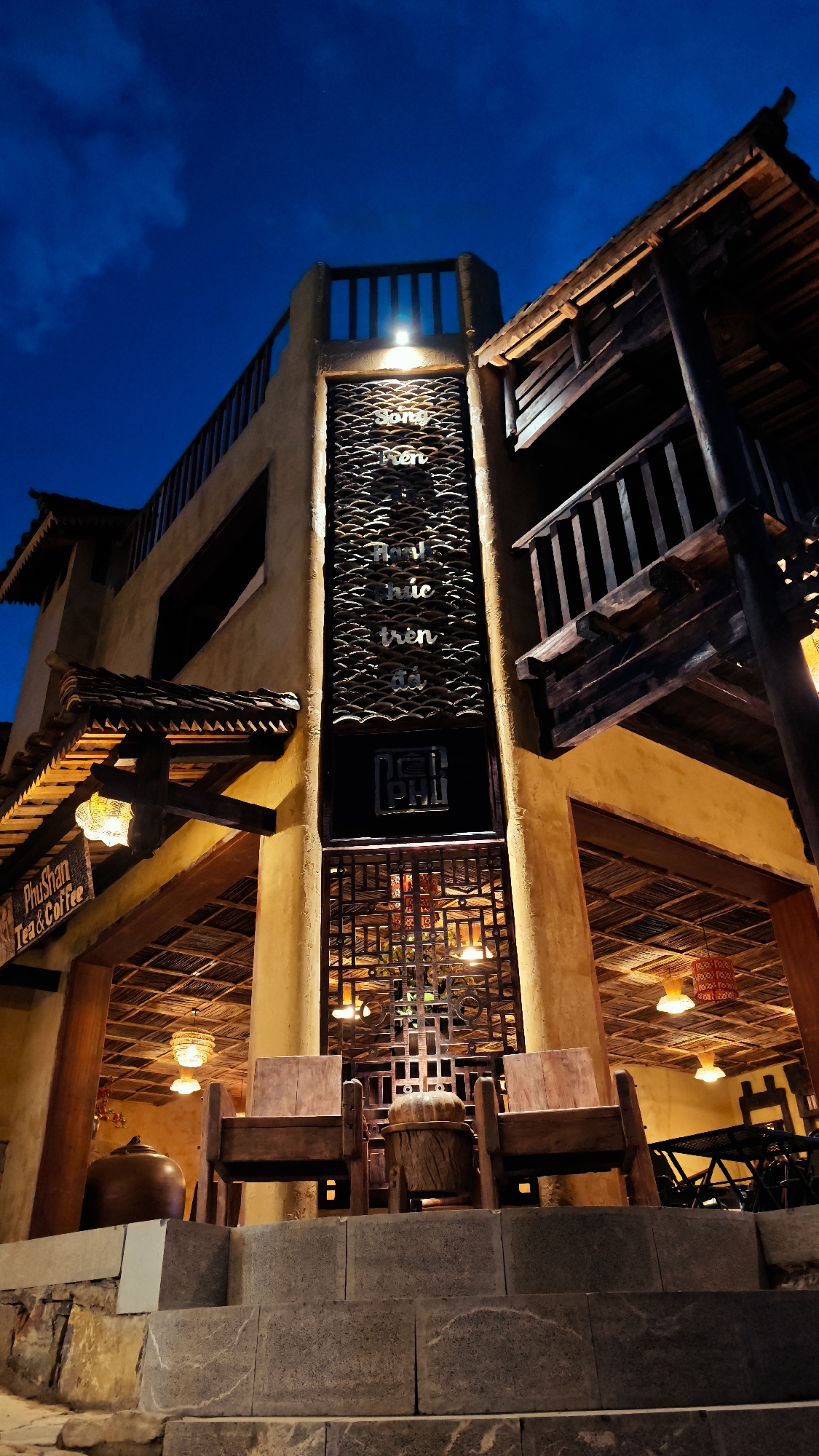 The image depicts the exterior of a building, seemingly a cafe or tea house called "PhuShan Tea & Coffee", located in Lũng Cú Commune, Vietnam. It is night, and the building is illuminated by internal lights and exterior fixtures. The architecture features a blend of traditional Vietnamese and modern styles. The building facade is primarily a light tan or yellow color, with wooden elements such as a balcony, roof eaves, and support beams. A tall, decorative screen runs vertically down the center of the building's front, displaying Vietnamese script and wave patterns. Below this screen, there is an enclosed, latticed structure. Two wooden chairs are positioned in front of the building on an elevated stone platform or steps. The interior, visible through the open front, has a lattice ceiling with hanging lamps. The sky above is a dark blue, indicative of twilight or night.