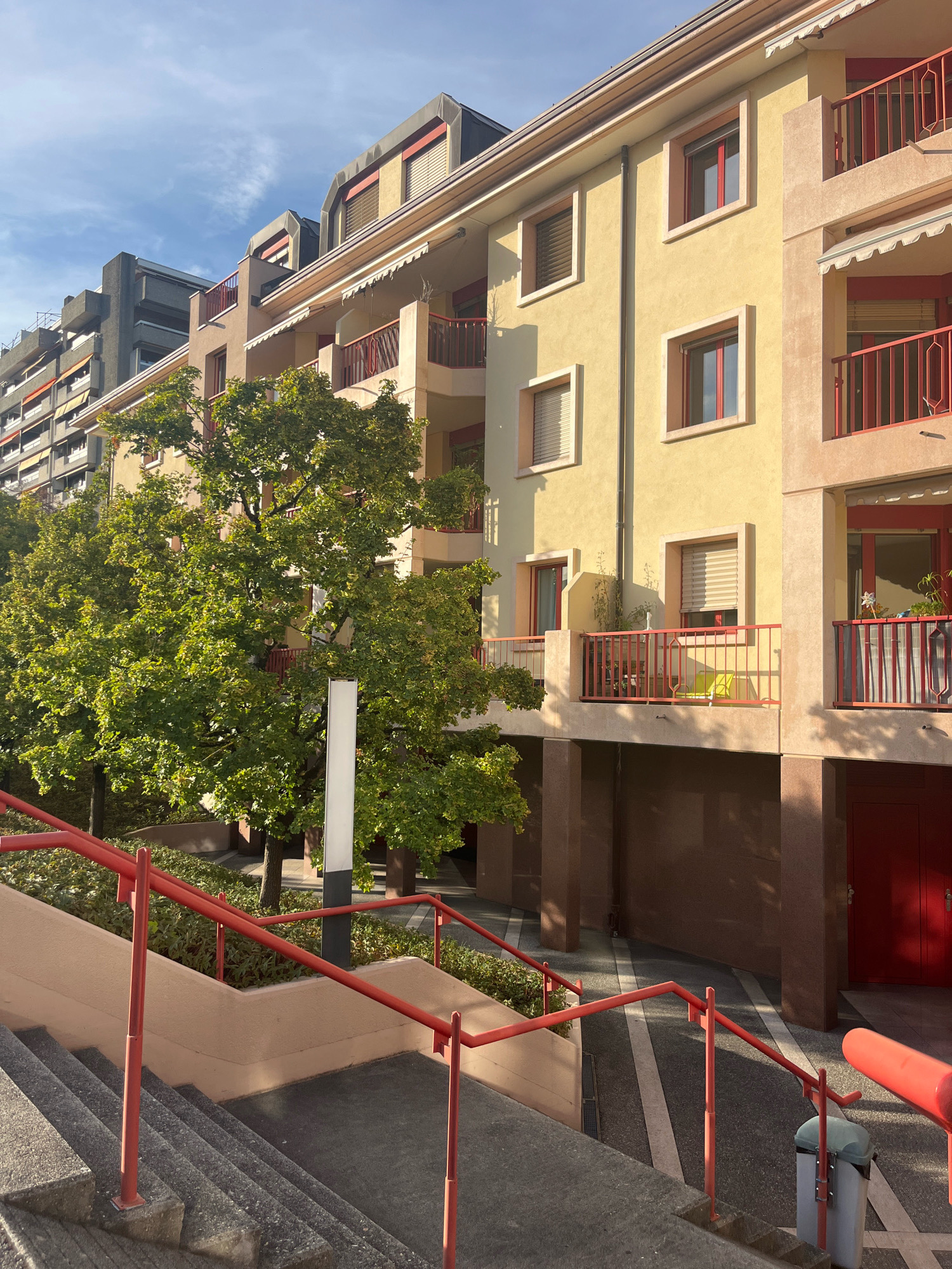 The image shows a close-up view of apartment buildings and a stairway with red railings in a city. The buildings have yellow and beige facades, with red balconies and windows. There is a large green tree in the foreground. The stairway is made of concrete and leads up towards the buildings. The scene appears to be an exterior shot, possibly taken on a sunny day, given the shadows cast by the buildings and tree. The style of the buildings suggests a European setting.
This is a residential building located at 23 Quai du Mont-Blanc, Geneva, Switzerland. The image captures the facade of the building, featuring its distinct yellow and beige coloration with red accents on the balconies and windows. A lush green tree stands prominently in front of the building, partially obscuring the lower floors. A concrete stairway with red railings is visible in the foreground, leading towards the entrance. The presence of awnings over some windows and balconies indicates a design adapted to the local climate. The architecture is typical of urban residential blocks found in Geneva.