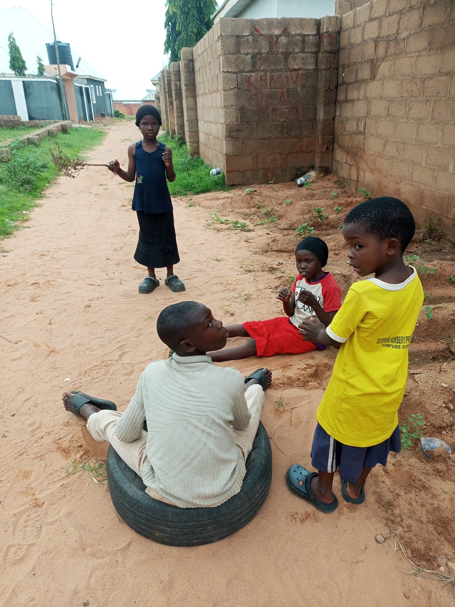 Four children are present in this outdoor scene, likely within the city of Tempure, Nigeria. The ground is covered in sandy dirt. In the foreground, a child wearing a light-colored, long-sleeved shirt and tan pants sits on a black tire, facing left and looking to the right. Their feet are clad in dark sandals. To the right of this child stands another child wearing a yellow t-shirt with writing on the back and dark blue shorts, also wearing dark sandals. This child is looking to the left. Behind them, a third child, wearing a red shirt and red shorts and a black head covering, is sitting on the ground facing right, holding something in their hands. In the background, a fourth child, wearing a dark blue sleeveless top and a black skirt, stands on a dirt path, holding a bundle of twigs and looking towards the viewer. The background features brick walls, a building with a dark roof, and some greenery.