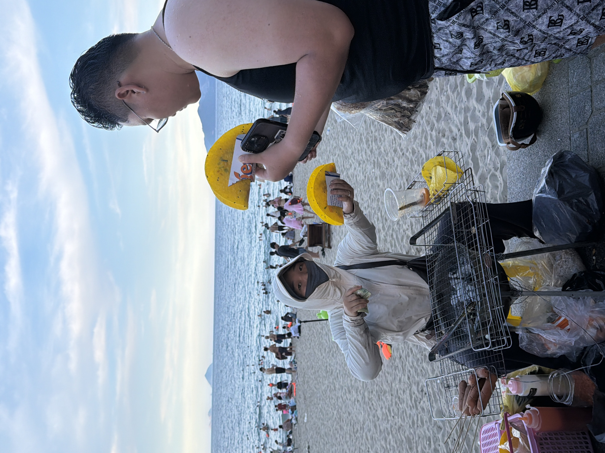 The image depicts a scene on a beach in Nha Trang Ward, Vietnam. The primary subjects are two individuals. The person in the foreground, wearing a light-colored hooded jacket and a face mask, is holding a handful of small items, possibly money, and looking towards the camera. This person is positioned on the sand, with the sea and numerous beachgoers visible in the background.
Behind and slightly to the left of the first person, another individual is leaning forward. This person is wearing a black tank top and glasses. They are holding up two yellow objects that appear to be flat, circular items, possibly snacks or decorative elements, with one of them also holding a smartphone.
The setting is a crowded beach with the ocean in the background under a bright, cloudy sky, suggesting daytime. The sand is visible in the foreground and across the beach. To the right of the first person, there is a collection of objects, including a wire rack containing food items like sausages on skewers, plastic bags filled with what looks like snacks, and a black trash bag. A helmet and other miscellaneous items are also visible near the ground. The overall impression is a busy beach environment with vendors or individuals selling items to beachgoers. There is no discernible text in the image.