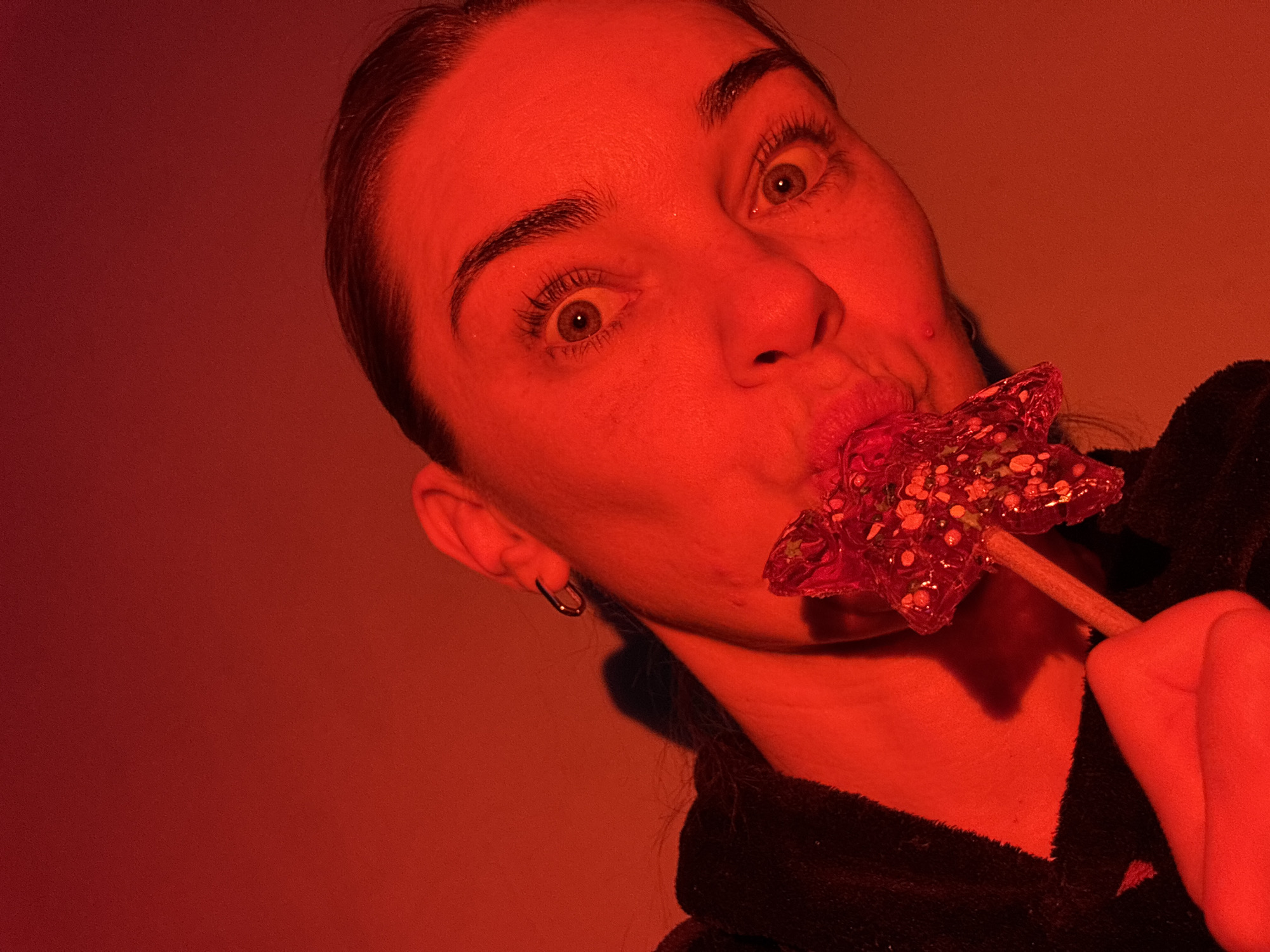 A woman with light skin and brown hair pulled back is illuminated by a reddish-orange light. She is holding a star-shaped lollipop to her mouth. She is wearing a black garment, possibly a robe or jacket. A hoop earring is visible in her left ear. The location and city of Unknown, Russia are not determinable from the image.
