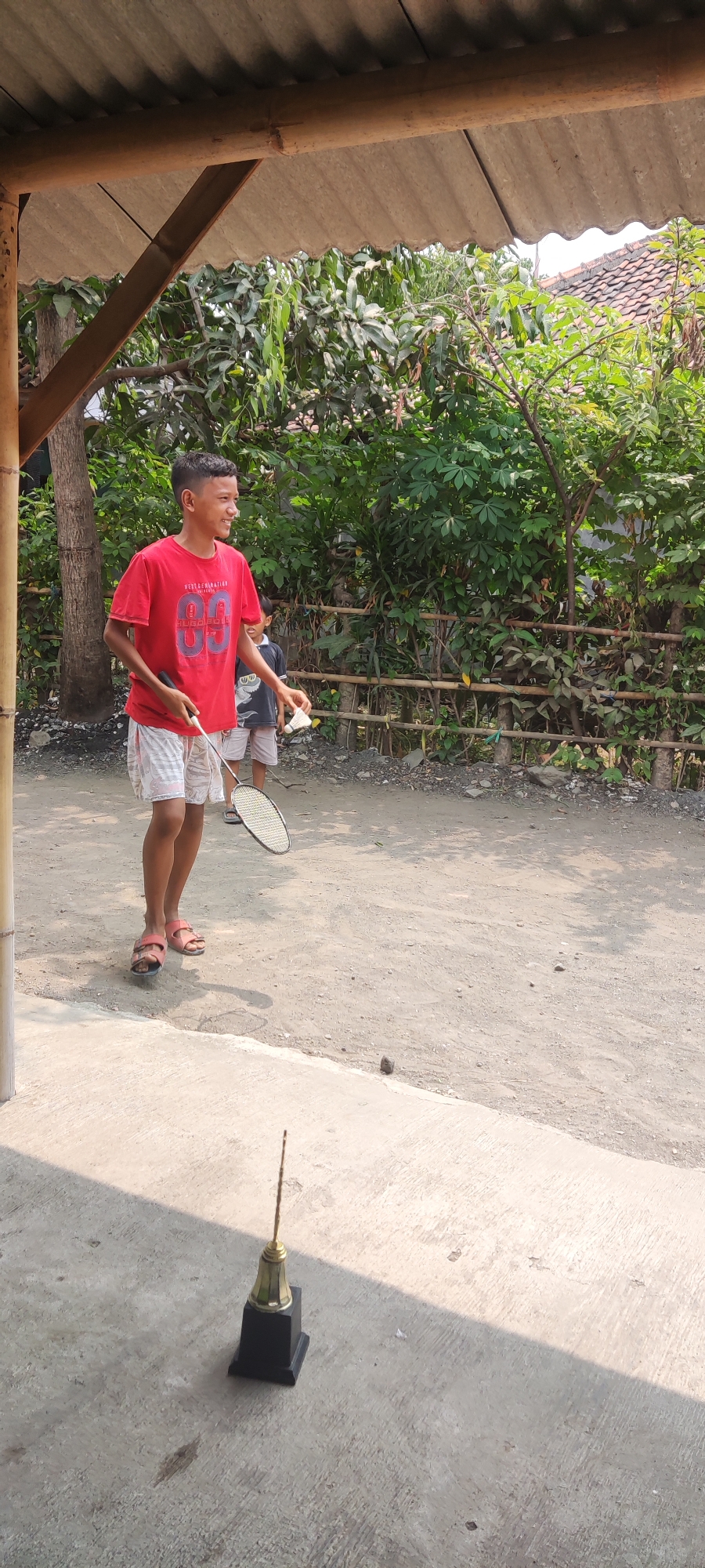 In Karangwangun, Indonesia, a young boy in a red t-shirt and checkered shorts stands outdoors, holding a badminton racket and a shuttlecock. He is positioned on a dirt ground under a makeshift shelter with a corrugated roof and bamboo supports. Behind him, a wooden fence borders lush green foliage. In the foreground, a black and gold trophy rests on a concrete surface. Another child, partially visible, stands near the fence. The scene appears to be a casual outdoor setting, possibly a yard or recreational area.