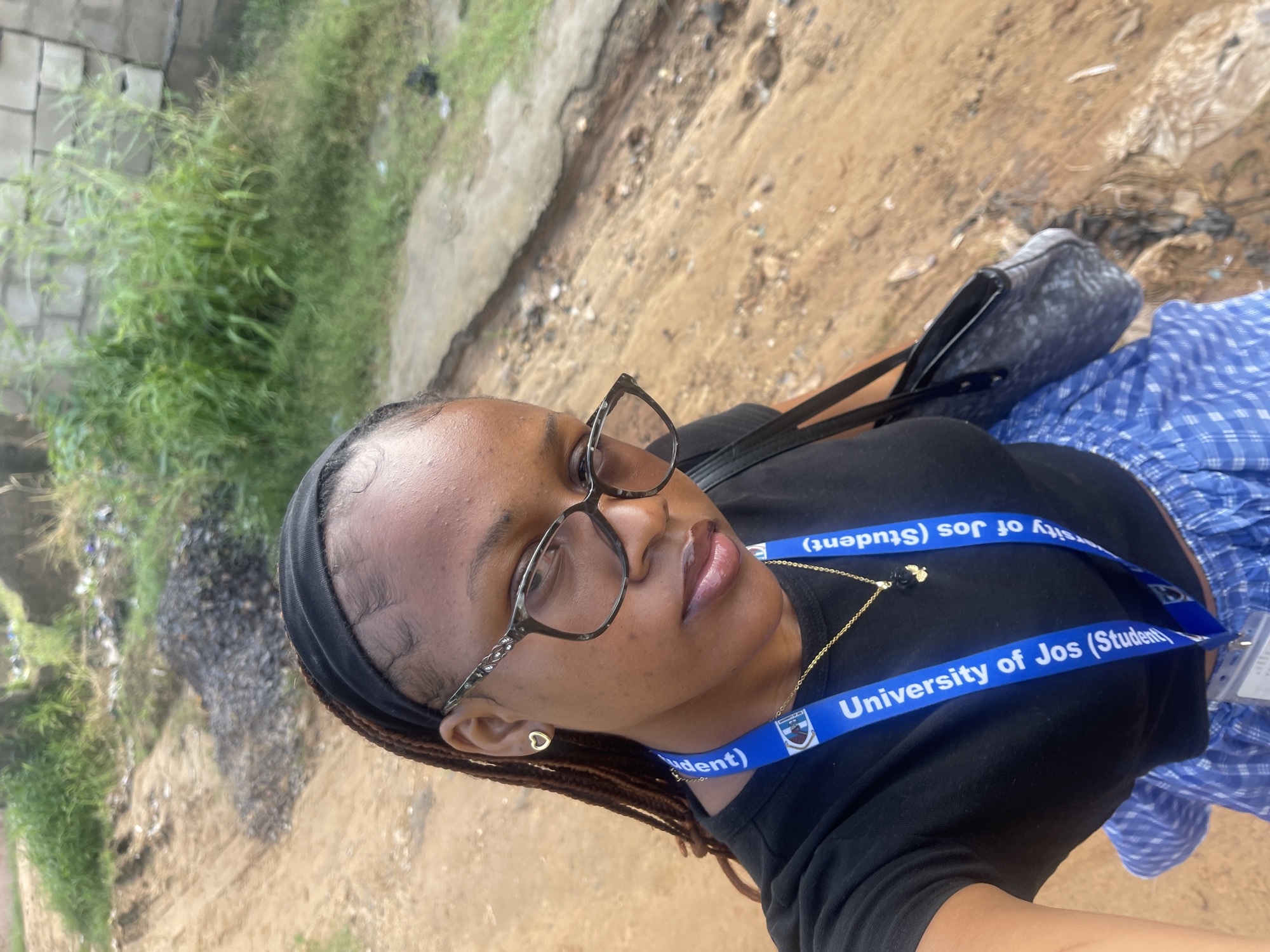 A medium shot shows a young Black woman with braided hair and a black headband, wearing glasses, a black t-shirt, and blue and white checkered shorts. She has a "University of Jos (Student)" lanyard around her neck and is holding a black handbag over her shoulder. The background appears to be an outdoor, unpaved area, possibly a construction or undeveloped site. The ground is sandy with some gravel and debris. In the upper right corner, there is a partially visible brick wall and overgrown vegetation. The overall impression is of an informal urban setting in Rafin Par, Nigeria.