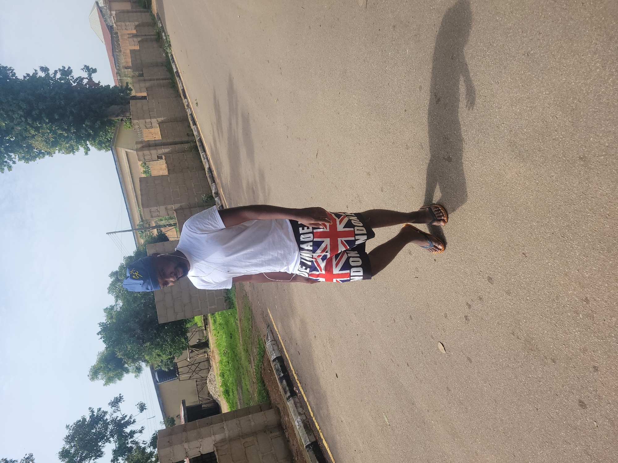 A man with dark skin and a short beard wears a white t-shirt, shorts with a Union Jack pattern and "LONDON" printed on them, and a blue cap with "SEI" printed on it. He is walking on a paved street with a curb and a yellow line. The background shows a brick wall, trees, buildings, and a water tower. The scene appears to be in Jos, Nigeria, based on the architecture and vegetation. The lighting suggests it is daytime, with a clear sky.