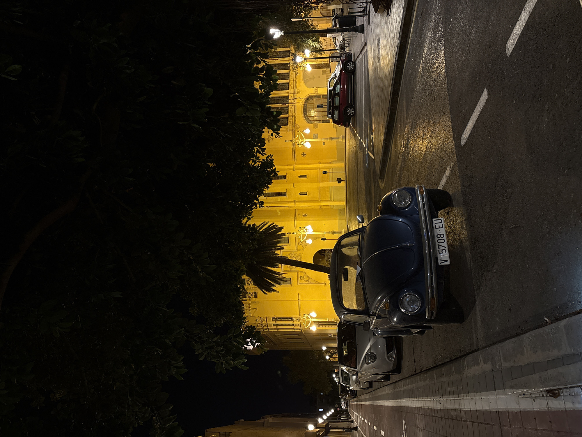 This is a nighttime photograph of a street scene in Valencia, Spain. In the foreground, a dark blue Volkswagen Beetle is parked at the curb. The car's license plate reads "V-5708-EU". To the left of the Beetle, a silver convertible is parked. Further down the street, several other cars are visible, all parked in a line. The street is paved with asphalt and marked with white lane dividers. A sidewalk lined with red and white tiles runs along the left side of the street.
In the background, a large, brightly lit yellow building stands out. The building has a classical architectural style with arched windows and ornate decorations. Several streetlights illuminate the scene, casting warm yellow light on the building and the wet pavement. Lush green trees with dark foliage overhang the street, creating shadows. One palm tree is visible between the Beetle and the yellow building. The overall atmosphere is quiet and serene, suggesting a late hour.