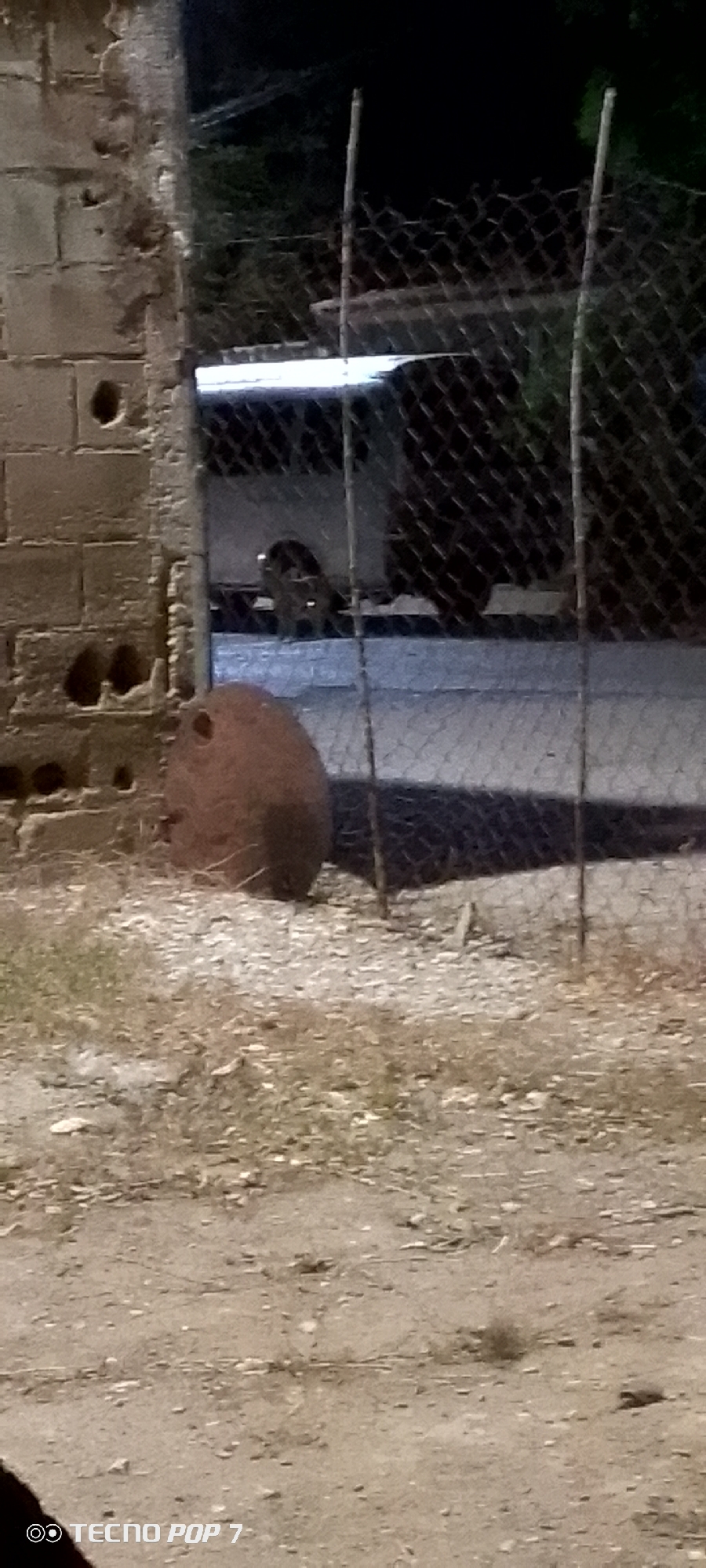 A partially obscured cylindrical object, possibly a tank or container, rests on sandy ground next to a dilapidated brick wall and chain-link fence. Behind the fence, a white vehicle is visible. The photo appears to be taken at night. The scene is likely located in a poorly maintained or industrial area. No people or interactions are visible in the image. The presence of the "TECNO POP 7" watermark suggests the photo was taken with a smartphone. No information indicates the city of Altagracia, Venezuela.