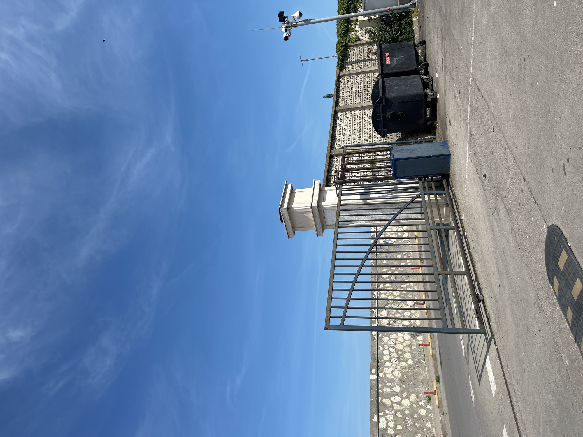This image captures an urban exterior scene in Varna, Bulgaria, under a clear blue sky with sparse, high-level cirrus clouds. The lighting suggests direct sunlight, casting distinct shadows.
In the foreground, a black speed bump with three yellow rectangular inserts lies on a grey asphalt surface. The asphalt is marked with a faded white line and displays minor scuffing and debris. A dark grey metal sliding gate, constructed from vertical bars with a curved top section, is open approximately one-third of its width, with its lower track and a single wheel assembly visible on the asphalt.
To the right of the gate, a rectangular blue utility box is positioned. Further right, against a textured concrete wall, stand two large, black, wheeled industrial waste bins. The rightmost bin has a red label displaying "SULO." The wall behind these bins is adorned with a repeating geometric pattern and partially covered with green ivy, particularly on the right side. Perched on top of this patterned wall, roughly centered in the midground, is a small, dark, bird-like figure. To the far right, a tall metal pole rises, supporting a cluster of security cameras and an antenna array.
To the left of the gate, a light-colored, multi-tiered rectangular structure, potentially a gatehouse or part of a building, is visible. Behind the open gate and to the left, a textured stone wall, possibly a retaining wall, is present, covered in various graffiti, including the letters "ABC" and "PGV." Beyond this wall, a paved road segment is delineated by a series of yellow curb segments and red bollards.
In the upper right quadrant of the sky, a small, dark, indistinct object, possibly a bird or insect, is visible in flight. No human subjects are present in the scene. The environment appears to be an access point or boundary for a property within an urban area.