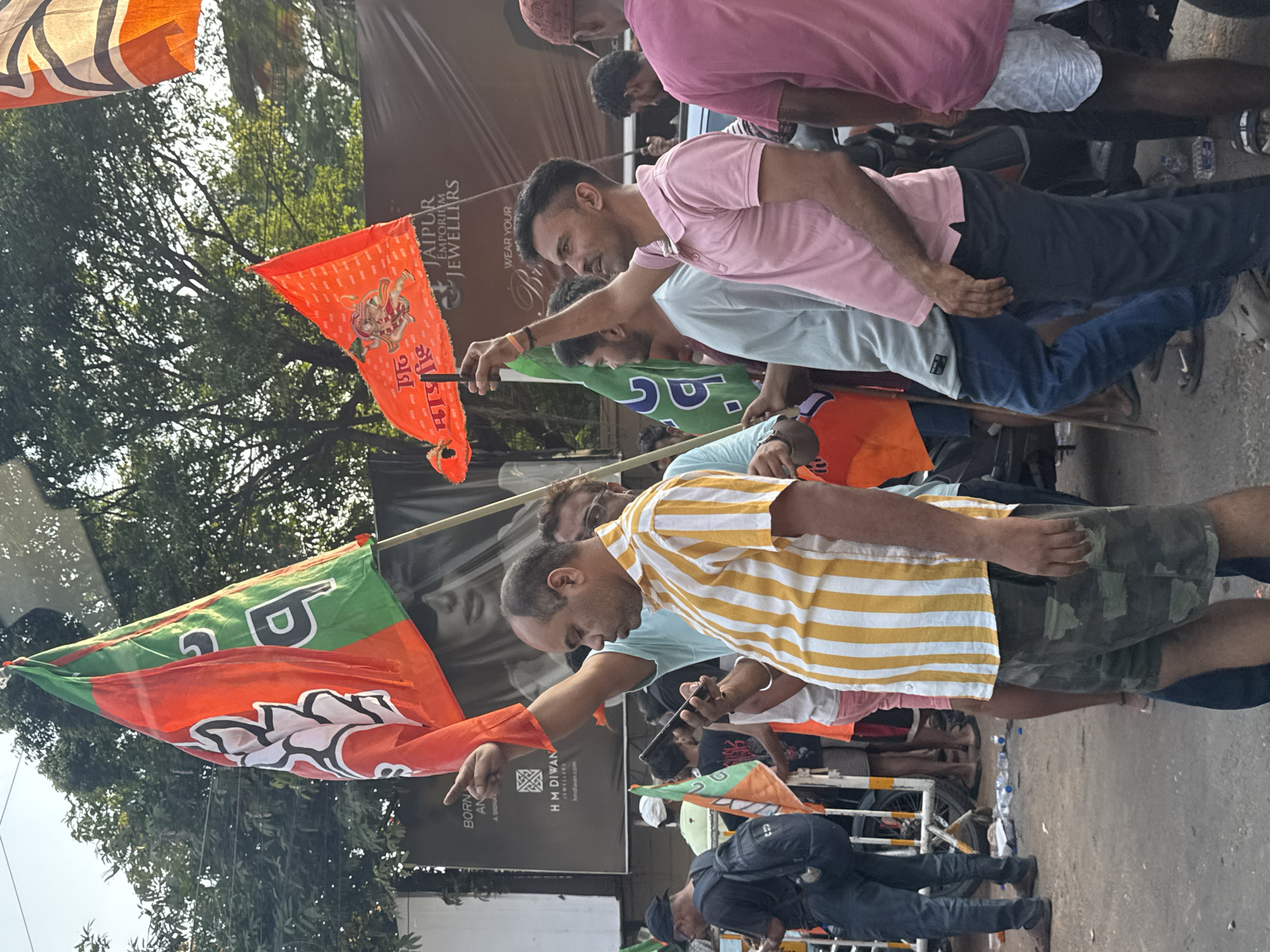 The image shows a political gathering in Kolkata, India. Several people are holding BJP (Bharatiya Janata Party) flags and chanting slogans. A man in a yellow striped shirt points toward something. The scene takes place near a Jaipur Emporium Jewellers store and H M DIWAN Jewellery store. There are other people in the background, including a policeman.
