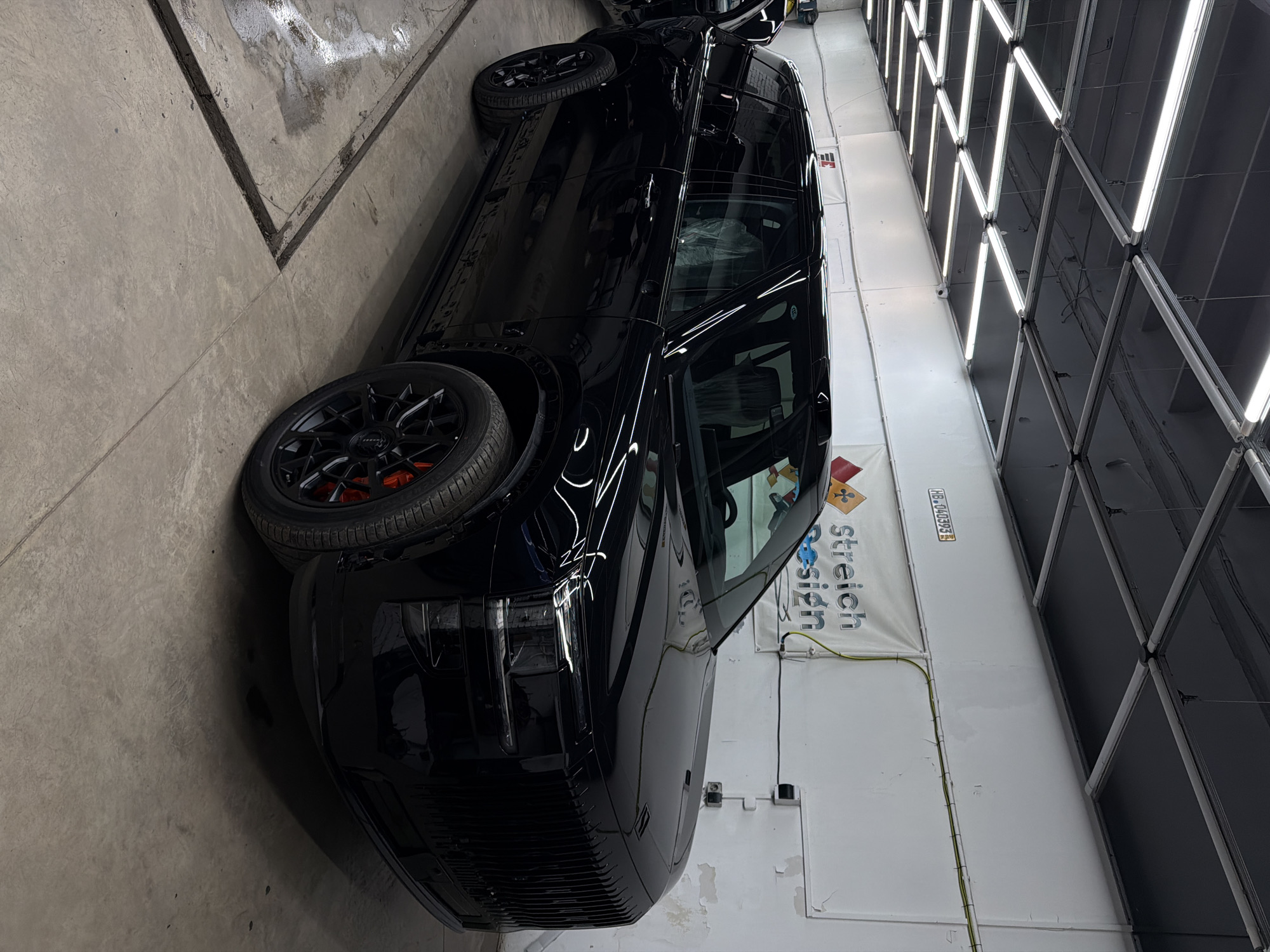 The image displays a sleek, highly polished black car, likely an SUV or large sedan, parked indoors in what appears to be a car detailing workshop or showroom located in Krasnoyarsk, Russia. The vehicle is positioned at an angle, showcasing its glossy bodywork, which strongly reflects the bright overhead lighting.
The car is equipped with black multi-spoke alloy wheels, beneath which vibrant red brake calipers are clearly visible. Its paint finish is exceptionally clean and reflective, suggesting a recent detailing or protective coating application. The windows are tinted dark, and some light-colored material, possibly protective seat covers or interior wrapping, can be seen through the back window.
The setting features a concrete floor that exhibits some wear and staining. The walls are white, with visible yellow and green electrical wires running horizontally across them. A prominent feature of the ceiling is a large grid of bright, linear LED lights arranged in a hexagonal or diamond pattern. These lights create striking, geometric reflections on the car's lustrous surface, emphasizing its pristine condition.
On the white wall to the right, a vertical white banner is partially visible, displaying the text "Stretch Design". Below this, a portion of another word is visible. Above the banner, a small white sticker or label bears the alphanumeric sequence "HB-040393 H". The scene conveys a professional automotive environment focused on vehicle aesthetics and care, with the bright artificial lighting ensuring optimal visibility for inspection. The time of day is not ascertainable from the indoor setting.