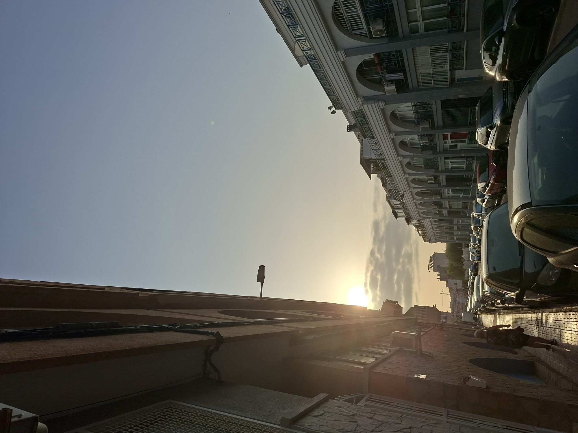 The image depicts a street scene in Torrevieja, Spain, during what appears to be sunset or early morning light, judging by the low angle of the sun and the warm glow. The perspective is from a street-level view, looking down a row of buildings and parked cars.
On the left side of the frame, a building with textured stone and stucco facades dominates the foreground, with arched entrances and what appear to be windows or alcoves visible. A person, walking away from the camera, is visible on the sidewalk in front of this building. They are wearing dark clothing. Exterior wiring and a lamp post are attached to the building.
The right side of the frame showcases a long row of multi-story buildings characterized by a consistent architectural style featuring a series of arched openings on each level, balconies with decorative railings, and visible air conditioning units. A line of parked cars, in various colors including black, silver, and red, fill the street in front of these buildings. The street itself is paved with interlocking bricks.
The sky is a pale blue, transitioning to a lighter hue near the horizon where the sun is visible. Scattered clouds are present, illuminated by the sunlight. In the distance, some taller buildings and construction cranes suggest a developing urban environment. The overall impression is a quiet residential or commercial street at a transitional time of day.