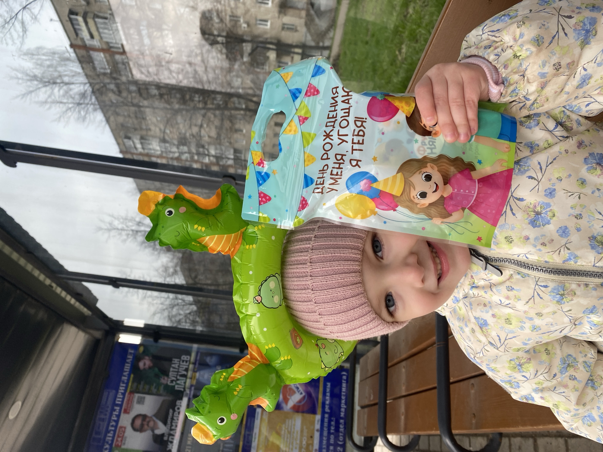 A young child, likely a girl, with blue eyes and a smiling expression, is pictured outdoors in what appears to be an urban setting in Chaikovsky, Russia. She wears a pink knitted beanie and a light-colored jacket with a delicate blue and green floral pattern.
The child is holding a colorful plastic gift bag with a cut-out handle. The bag features illustrations of a cartoon girl with balloons and party hats. Prominently displayed text on the bag, in Cyrillic, reads: "ДЕНЬ РОЖДЕНИЯ У МЕНЯ, УГОЩАЮ Я ТЕБЯ!" which translates to "My birthday, I treat you!"
An inflatable green dragon toy with orange accents is placed on or near her head, contributing to a playful atmosphere. The child is seated on a wooden bench with dark metal supports.
The background shows reflections of a tall building with numerous windows and bare tree branches, indicative of late winter or early spring, under an overcast sky. Behind the child and to her left, advertisements are visible on what appears to be a bus stop or public information panel. Visible Cyrillic text includes "КУЛЬТУРЫ ПРИГЛАШАЕТ" (Culture invites), names such as "СУЛТАН ЛАГУЧЕВ" and "ЖЛАМИРОВ", a date "24.05", a number "700", and phrases like "размещения рекламы" (ad placement) and "отдел маркетинга" (marketing department). The overall mood is cheerful, likely capturing a moment during a birthday celebration or gift exchange.