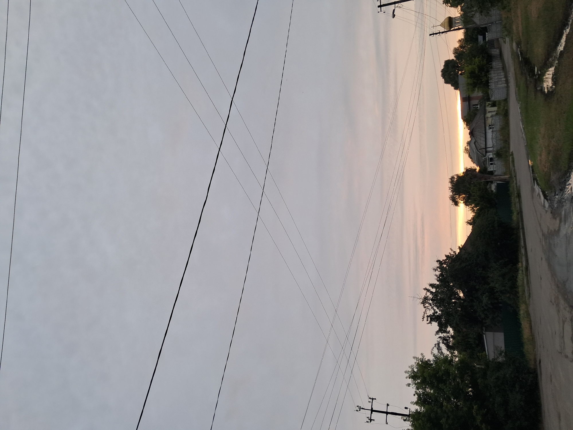 The image captures an overcast sky over a rural or suburban setting in Kremenchuk, Ukraine, likely during twilight due to the soft, diffused light and the warm glow on the horizon. Numerous power lines crisscross the sky, originating from utility poles visible on the left and right sides of the frame. Below the sky and power lines, a road curves through a scene populated by trees and buildings. A power pole with insulators stands on the left. On the right, a golden-domed church is partially visible behind trees. The road appears to be paved but shows signs of wear and a water-filled rut. Houses with fences are scattered throughout the middle ground, surrounded by dense foliage. The overall impression is one of a quiet, somewhat undeveloped residential area under a muted sky.
