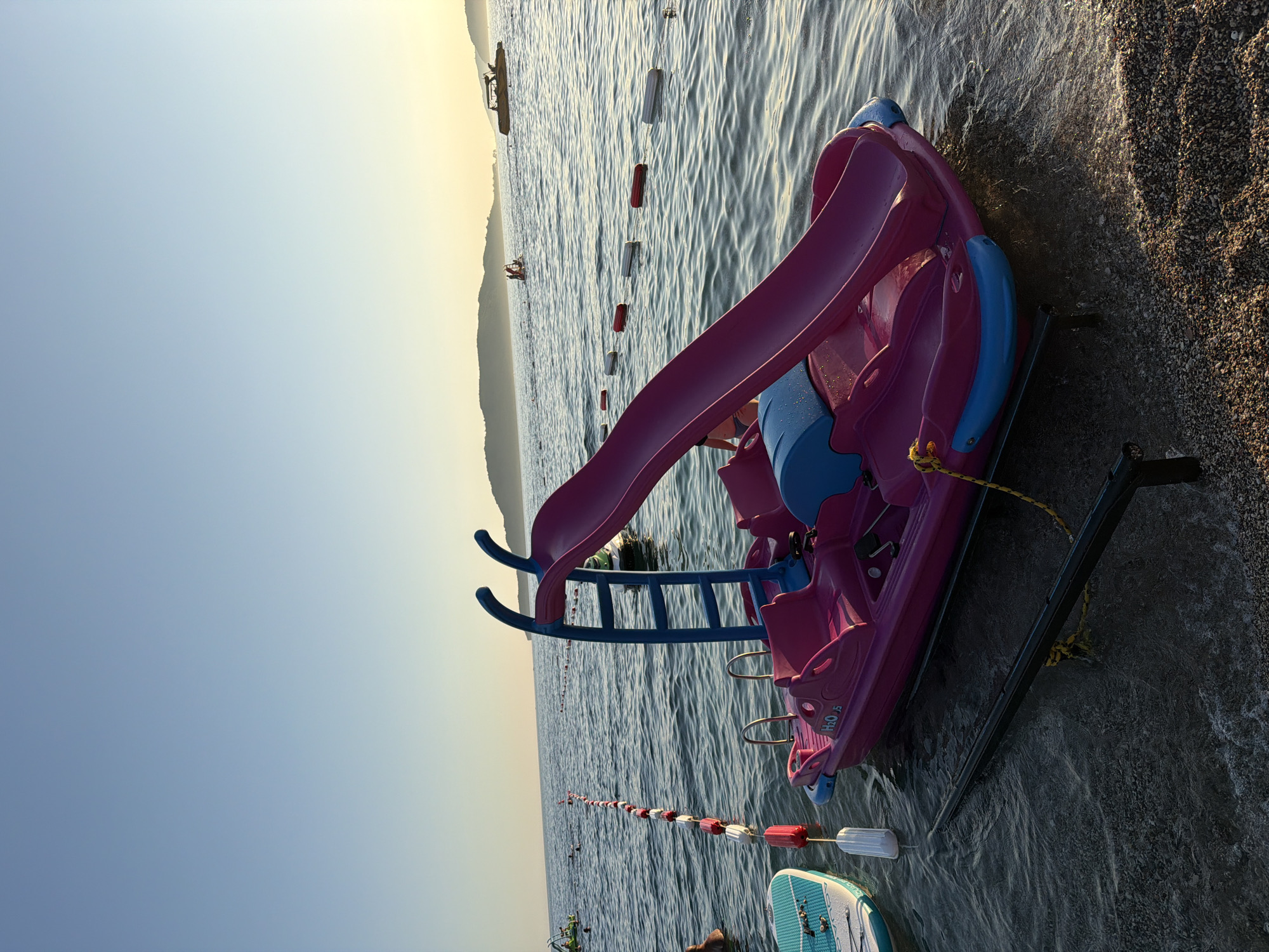 A pink and blue pedal boat with a slide sits partially submerged in the shallow water of Boreti beach in Montenegro, with a clear sky and calm sea. A white and blue paddleboard is to the left of the pedal boat. A rope with red and white buoys stretches across the water, separating a swimming area. In the background, several people are visible in the water, and a jet ski is seen further out at sea. Hills line the horizon, bathed in the warm light of what appears to be early morning or late afternoon. The beach is composed of small pebbles and sand.