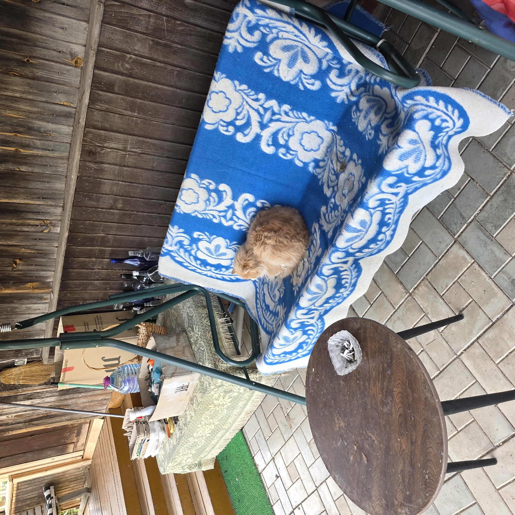The image depicts an outdoor setting in Koshelevichi, Russia. A wooden structure, possibly a porch or shed, is visible with vertical wood paneling. A metal swing bench with a blue and white patterned blanket is positioned on a paved patio. An orange cat is curled up on the blanket. A round wooden table with black metal legs stands nearby, holding a plastic bag containing cigarette butts. Various items are cluttered around the base of the swing, including a water bottle, a broom, and cardboard boxes. The ground is covered in grey paving stones.