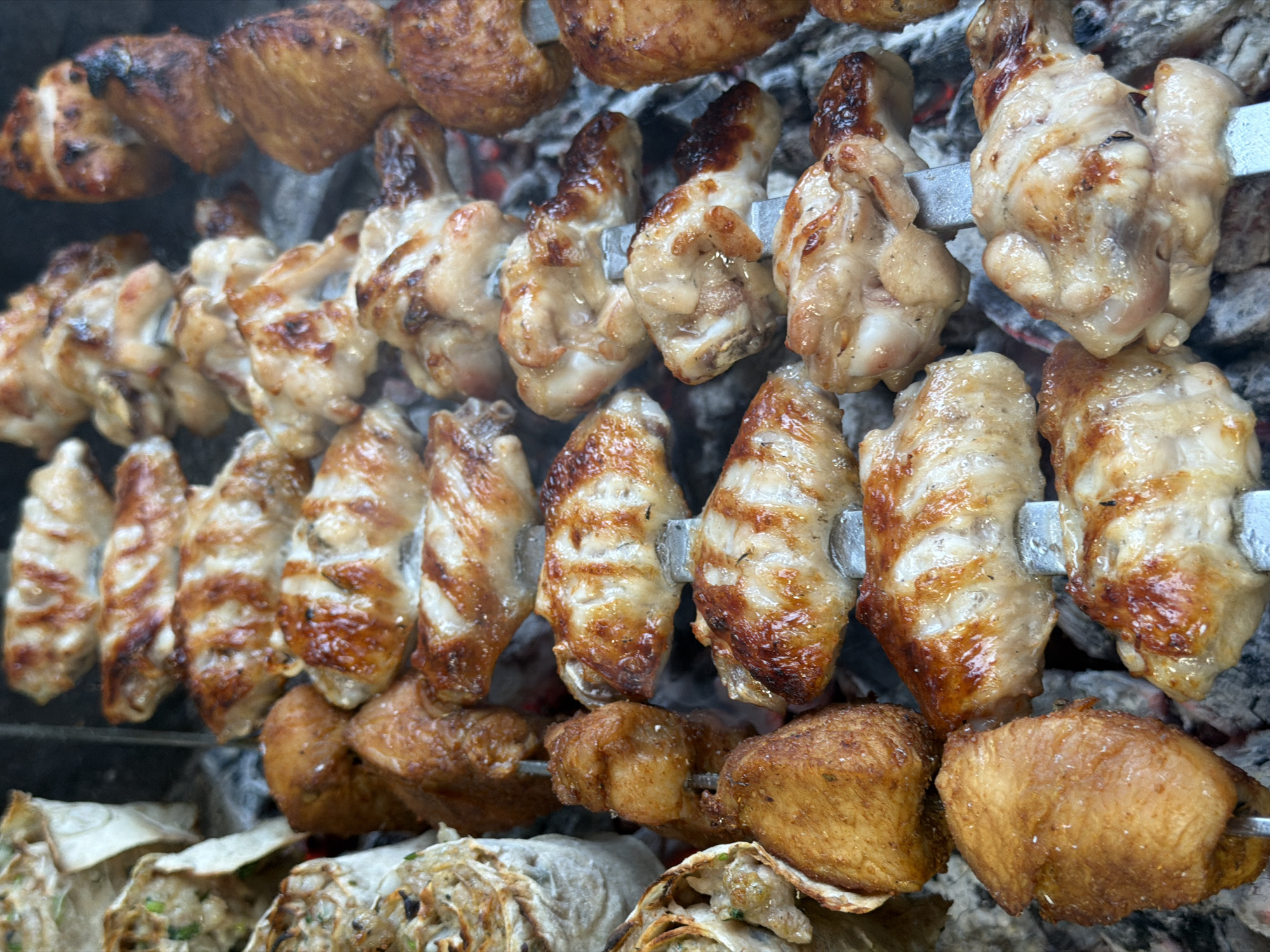 A close-up, high-angle shot shows chicken wings and what appear to be rolled flatbreads with filling, cooking on a charcoal grill. The chicken wings are skewered and have grill marks, some with slightly charred edges. The flatbreads are golden brown and appear to be layered. The scene is set in Tuapse, Russia, as indicated by metadata. The lighting is natural and appears to be daylight. The overall impression is of a casual outdoor cooking event.