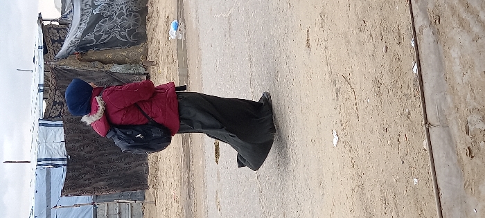 An individual, likely female, is depicted walking from right to left on a dirt and rough asphalt path in Khan Yunis, Palestinian Territory. The person wears a dark blue head covering, a red puffer jacket with a fur-lined hood, a long black skirt, and dark shoes, carrying a black backpack. Their right foot is slightly lifted, indicating motion. In the background, multiple makeshift shelters are constructed from white tarpaulins, dark patterned fabrics, and lighter sheets, supported by poles on sandy ground. The sky is overcast. The path features visible tire tracks, small depressions, and scattered debris including a blue plastic bag and white scraps. A low concrete curb-like structure runs across the lower foreground.
