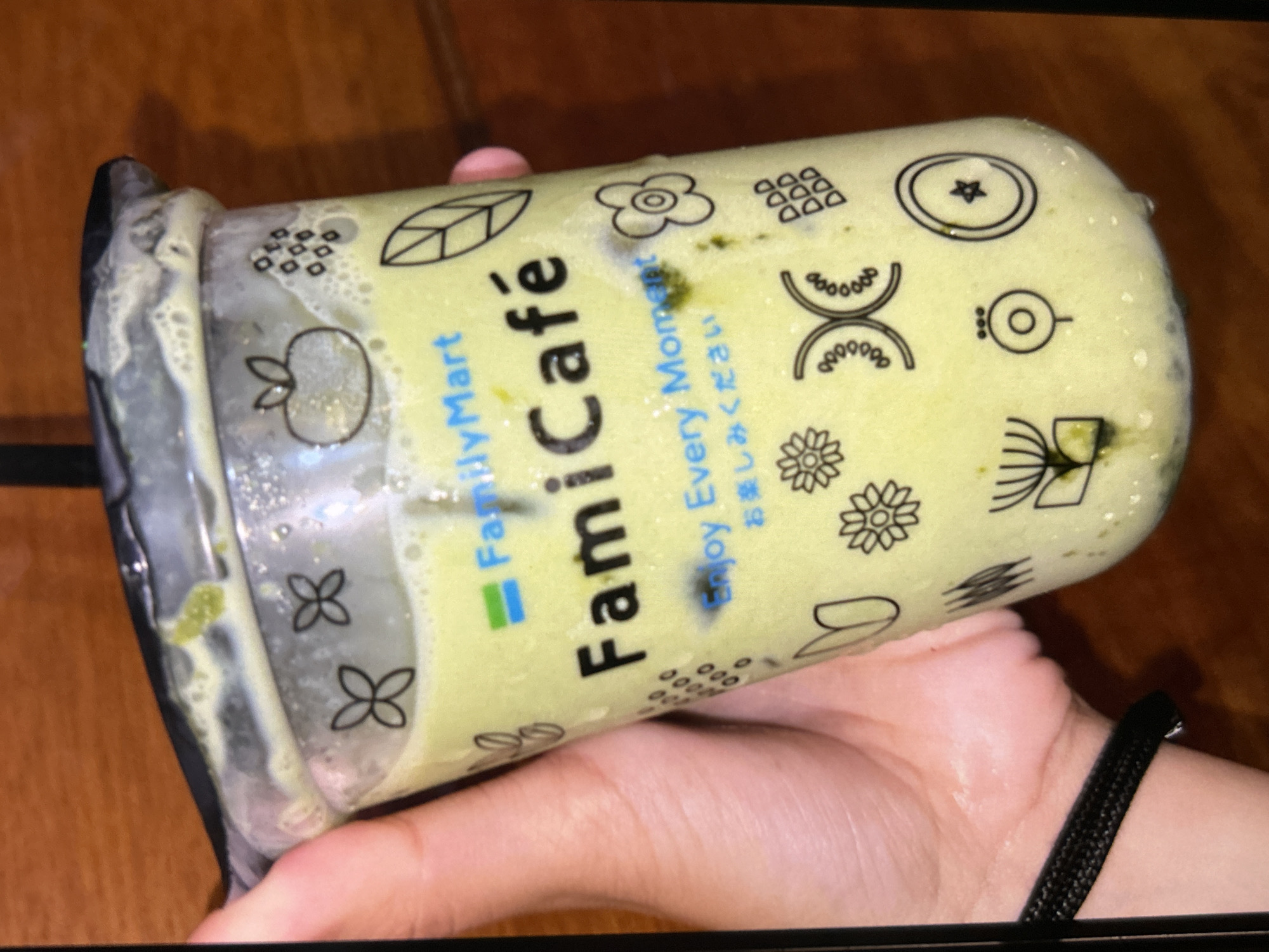 A hand holds a clear plastic cup filled with a light green beverage, likely a smoothie or iced coffee drink. The cup is branded with "FamilyMart" and "FamiCafé" in white and black lettering. Decorative black line drawings of flowers, leaves, and abstract shapes are printed on the cup's exterior. The beverage appears frothy and contains some solid green particles. The background consists of a wooden surface, suggesting the photo was taken indoors or on a table. No specific location in Depok, Indonesia is identifiable from the image.