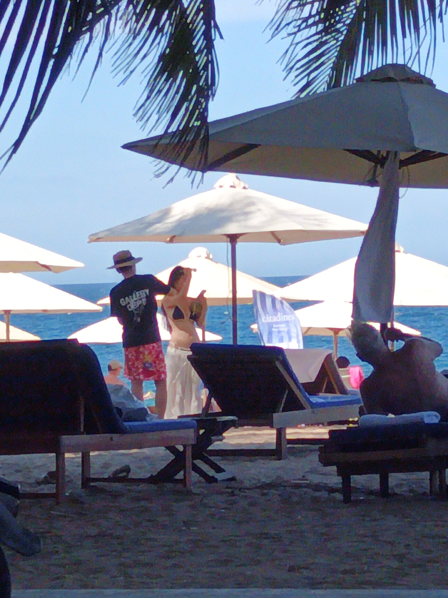 The image depicts a beach scene, likely at a resort in Nha Trang, Vietnam. In the foreground, several dark-colored lounge chairs are visible, with one in the lower left partially obscured. The sand appears light brown and grainy.
In the mid-ground, a man wearing a straw hat, dark t-shirt with "GALLERY DEPT" printed on the back, and red and pink patterned shorts stands facing away from the viewer. He is holding a coconut. To his right, a woman in a bikini top and a long white skirt is partially visible, also holding a coconut. They are standing near beach umbrellas, one of which is large and dark gray, and another is white. Several other white beach umbrellas are scattered further in the background.
The ocean is visible behind the people and umbrellas, with calm blue water. A blue and white striped flag with the word "citadines" is planted in the sand to the left of center. The sky is clear and blue. In the far background, the silhouette of a person is visible reclining on a lounge chair. The scene suggests a relaxed atmosphere on a sunny day at the beach.
