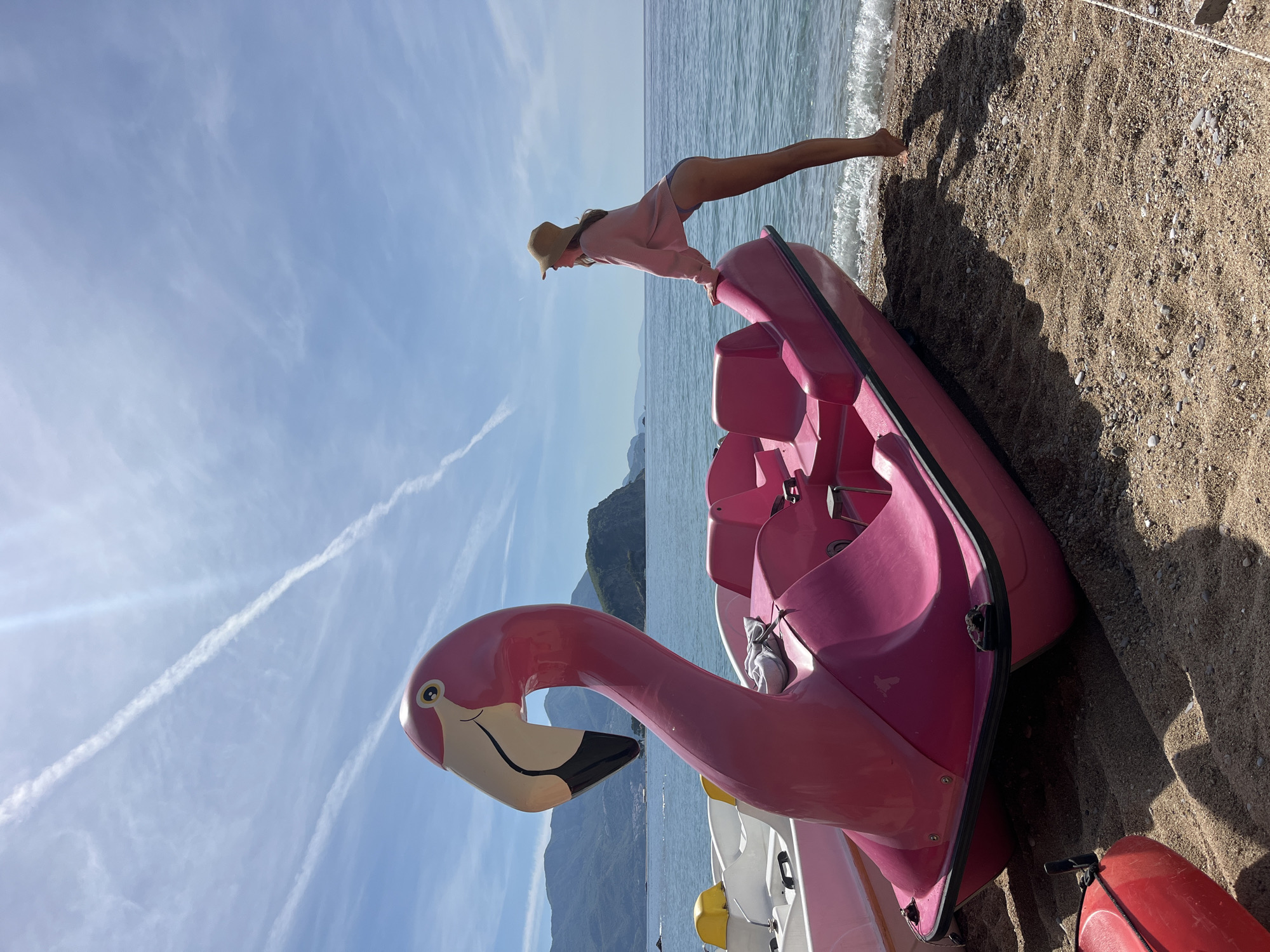 The image shows a pink flamingo-shaped paddle boat on a sandy beach in Budva, Montenegro. A woman in a sun hat, pink sweater, and swimsuit is leaning over the boat, her feet touching the water's edge. In the background are mountains and a blue sky with contrails. Other paddle boats are visible beneath the pink one. The beach is light brown.