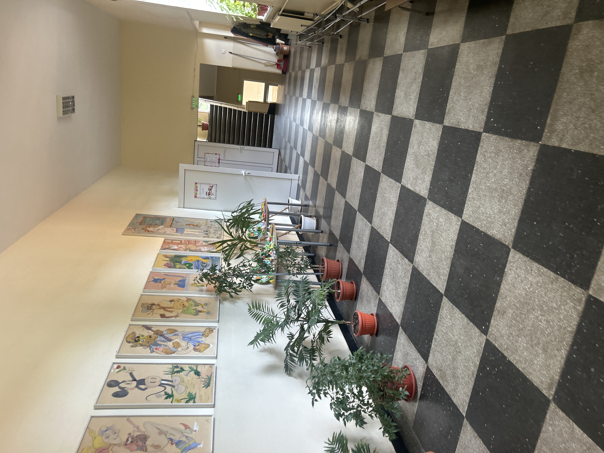 The image shows a school hallway in Loznitsa, Bulgaria. The hallway has a checkerboard floor pattern of black and gray tiles. Along the left wall, there is a series of framed children's drawings featuring cartoon characters like Mickey Mouse and Alf. Several potted plants are placed along the wall and in front of a table displaying more artwork and craft projects.
On the right side of the hallway, there is a set of metal chairs stacked against the wall. Further down the hallway, a staircase leads to an upper level, with a white door to the left of the stairs. A broom and dustpan are visible near the stairs, suggesting cleaning activities. Several coats are hung on a rack near a window. The overall impression is of a well-used, colorful, and decorated educational space.