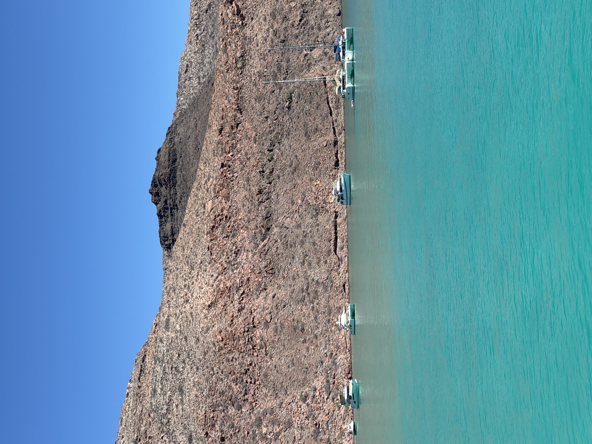 The image depicts a rocky cliff face on the left side, contrasting with a body of turquoise water on the right. Several boats are docked along the edge where the cliff meets the water. The sky is a clear blue above the cliff. The location is Unknown, Mexico.