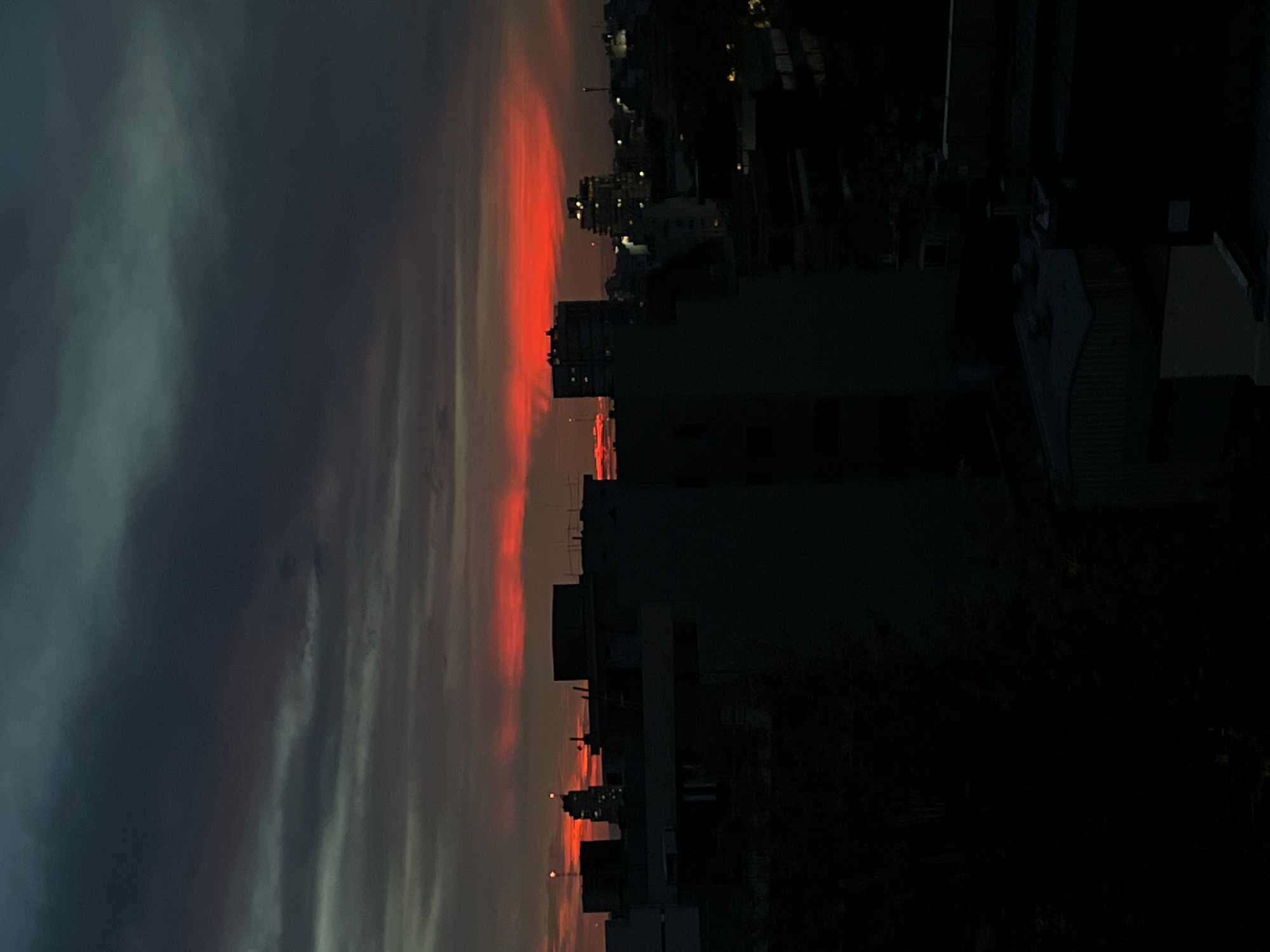 A low-angle, long shot depicts a cityscape at dusk in Buenos Aires, Argentina. The buildings are silhouetted against the sky, which features a striking sunset. Streaks of orange and red are prominent near the horizon. To the left, the sky transitions to darker gray and blue hues. The buildings are mostly dark and indistinct, although some lights can be seen in windows. The image appears to be taken from an elevated position, offering a view over the urban landscape. There is no human activity visible.