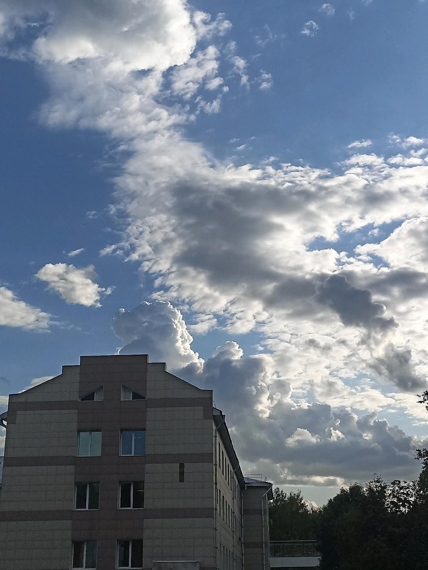 The image shows a modern building in Novosibirsk, Russia, under a dramatic sky filled with cumulonimbus clouds. The building appears to be a multi-story structure with a facade of light-colored tiles and horizontal bands of darker tile. It has multiple windows, some reflecting the bright sky. The roofline is angular, with triangular elements. Trees and other foliage are visible in the lower right and middle ground, suggesting an urban park or natural area adjacent to the building. The sky dominates the upper portion of the image, with a mix of bright white and darker gray clouds, indicating dynamic weather conditions. There are no people visible in the scene. The lighting suggests it is daytime, possibly late afternoon, given the intensity of the sunlight illuminating the clouds. The image captures a sense of an impending weather change or a dramatic sky event.