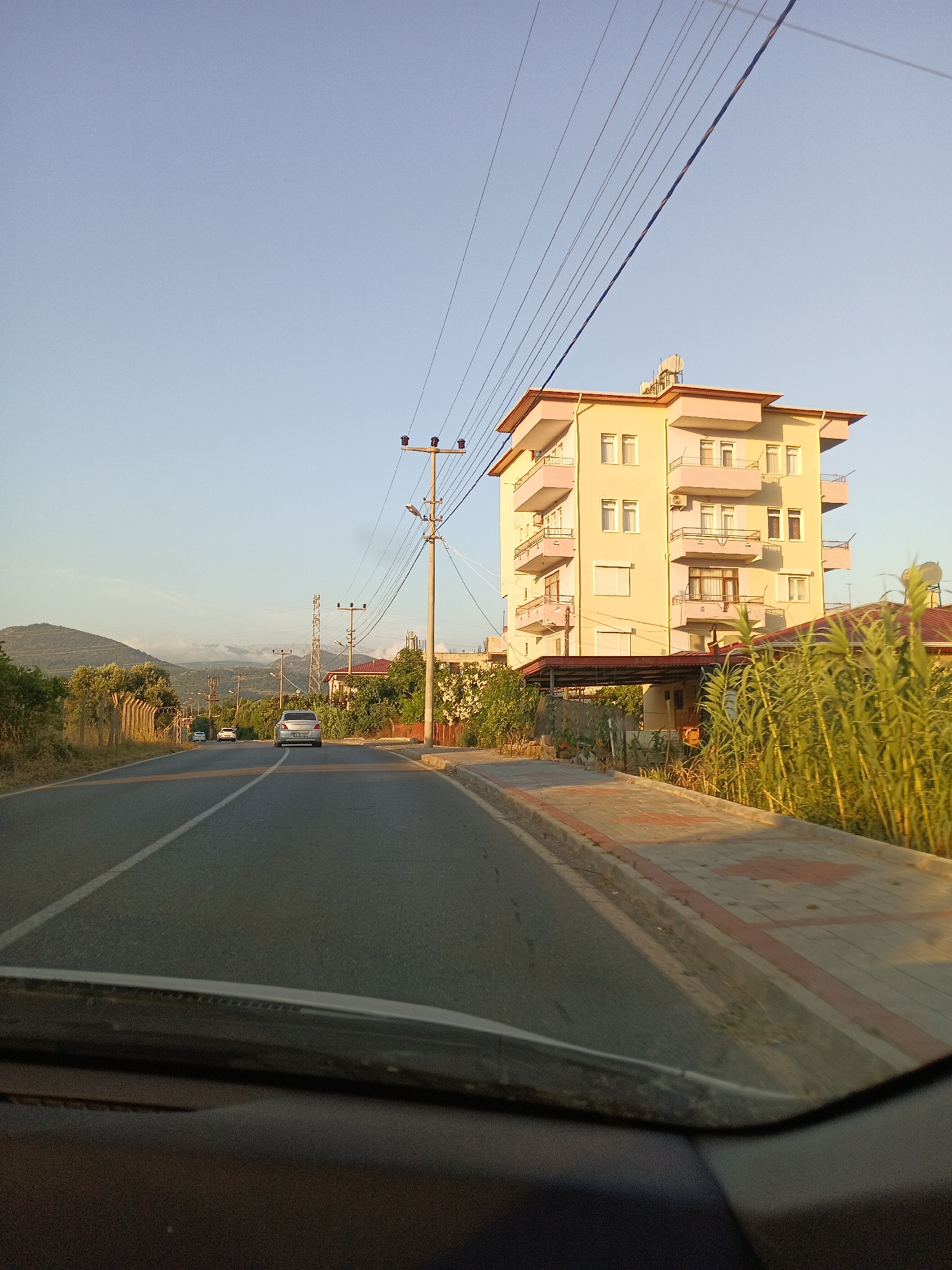 The image captures a street scene in Gazipaşa, Turkey, under a clear blue sky. The perspective is from inside a vehicle, looking forward. A paved road with a white lane divider stretches into the distance. To the right of the road is a sidewalk with red and grey patterned pavers, bordered by a curb. Beyond the sidewalk, a multi-story yellow building with pink trim stands prominently. It has several balconies and windows, and air conditioning units are visible. Tall green reeds grow along the edge of the property next to the building. Power lines crisscross the sky above. Further down the road, several cars are visible, and on the left side, there are trees and a wire fence. In the background, hills rise, with some clouds gathered near their peaks. The lighting suggests it is late afternoon or early evening.