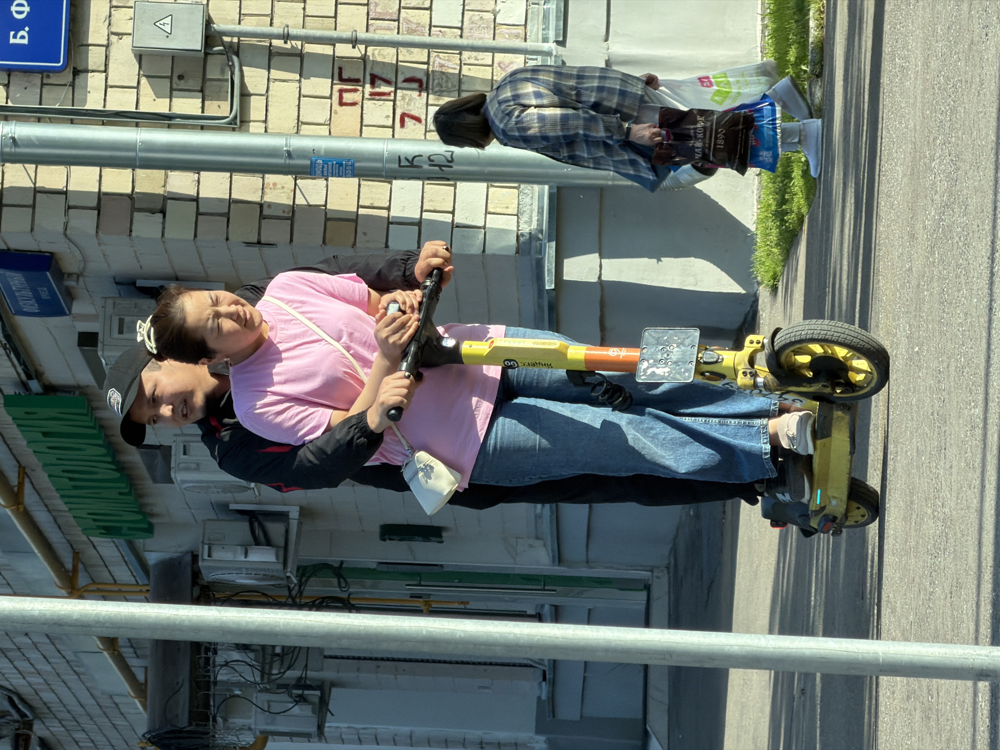 Two individuals, a man and a woman, are riding an electric scooter down a paved sidewalk in Moscow, Russia. The man is positioned behind the woman, with his hands on the handlebars, and his body obscuring most of her back. The woman is in front, her hands also on the handlebars. She is wearing a pink t-shirt and blue jeans, and has a white purse slung across her body. The man is wearing a black jacket and a black baseball cap. The scooter is yellow and white.
In the background, a building with light-colored brickwork is visible. A green sign with Cyrillic lettering is mounted on the building. There are also air conditioning units mounted on the exterior wall. A street pole with signage is also present. Further in the background, another woman is walking, carrying shopping bags. The scene is illuminated by sunlight, casting shadows on the ground. The overall setting appears to be an urban environment, likely a residential or commercial street in Moscow.