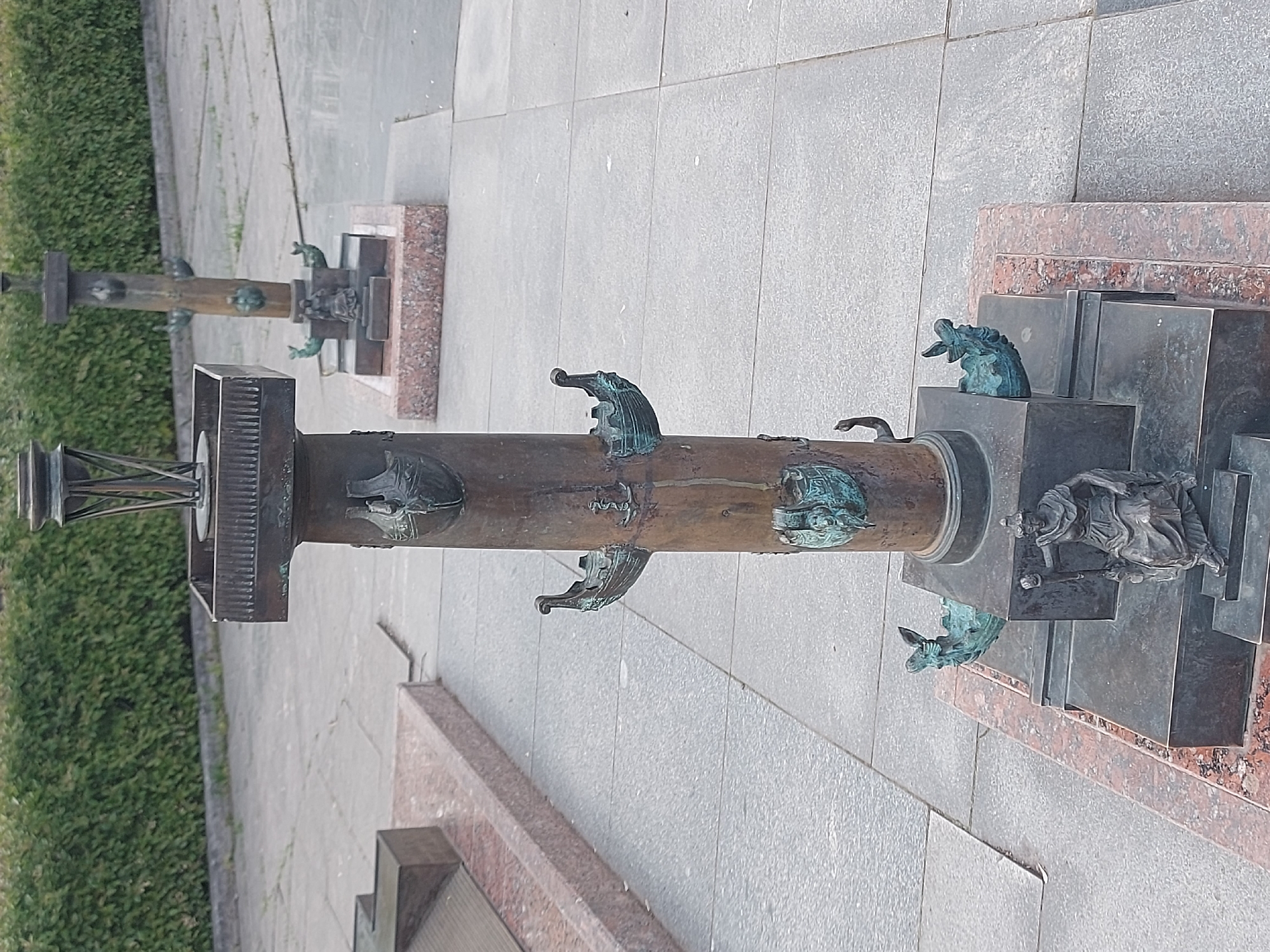 The image shows a pair of Rostral Columns in Saint Petersburg, Russia. The columns are made of dark metal and are adorned with bronze prows and figureheads of ships. The columns sit on dark gray pedestals. The surrounding pavement is light gray, with patches of grass visible.