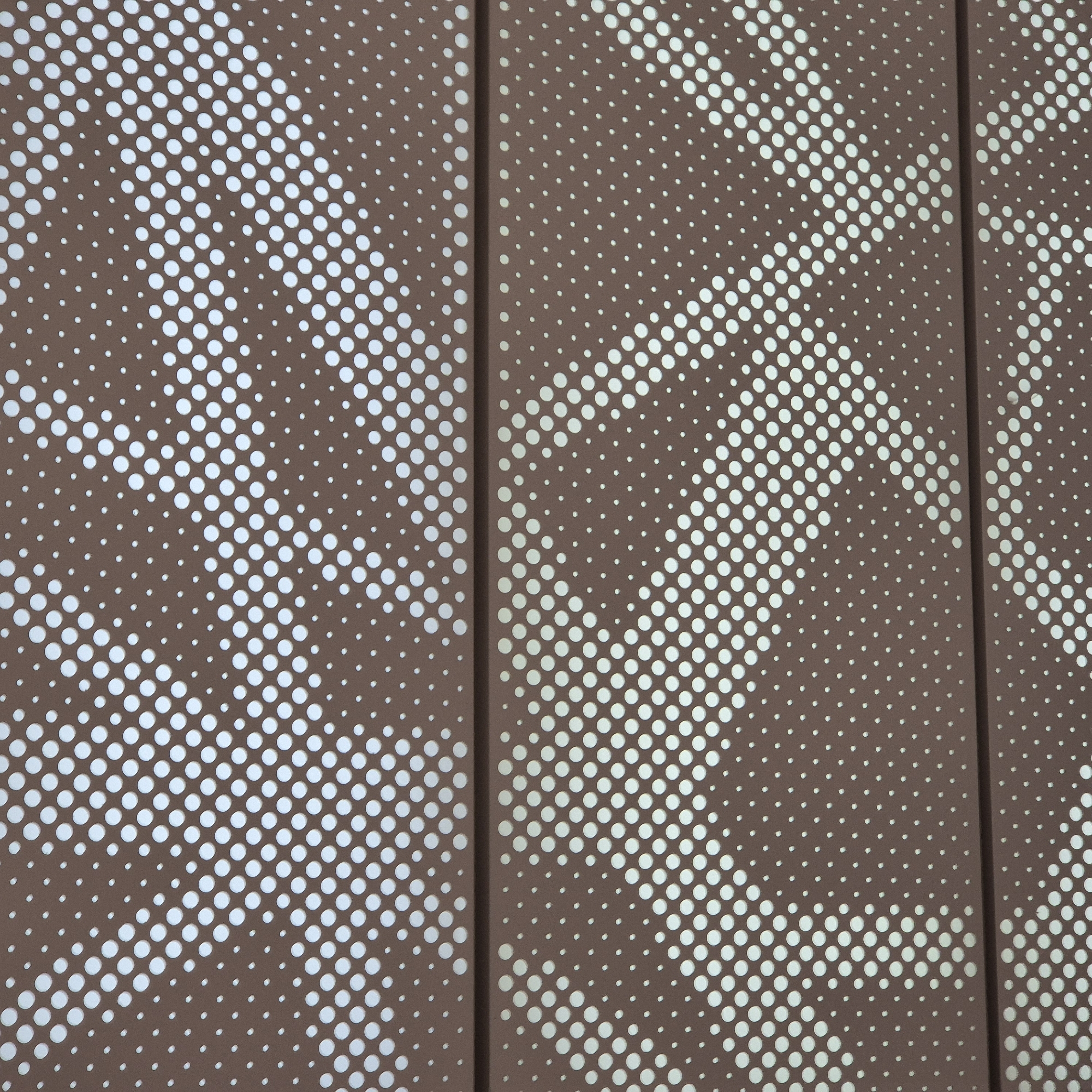 The image shows a close-up view of a brown perforated metal panel with a pattern of white dots of varying sizes. The dots are arranged in diagonal rows, with larger dots closer together and smaller dots further apart, creating a gradient effect. There is a horizontal seam across the middle of the image, dividing the panel into two sections. The background visible through the perforations is light blue. The scene appears to be an outdoor architectural feature, possibly a facade or railing, in an urban setting.