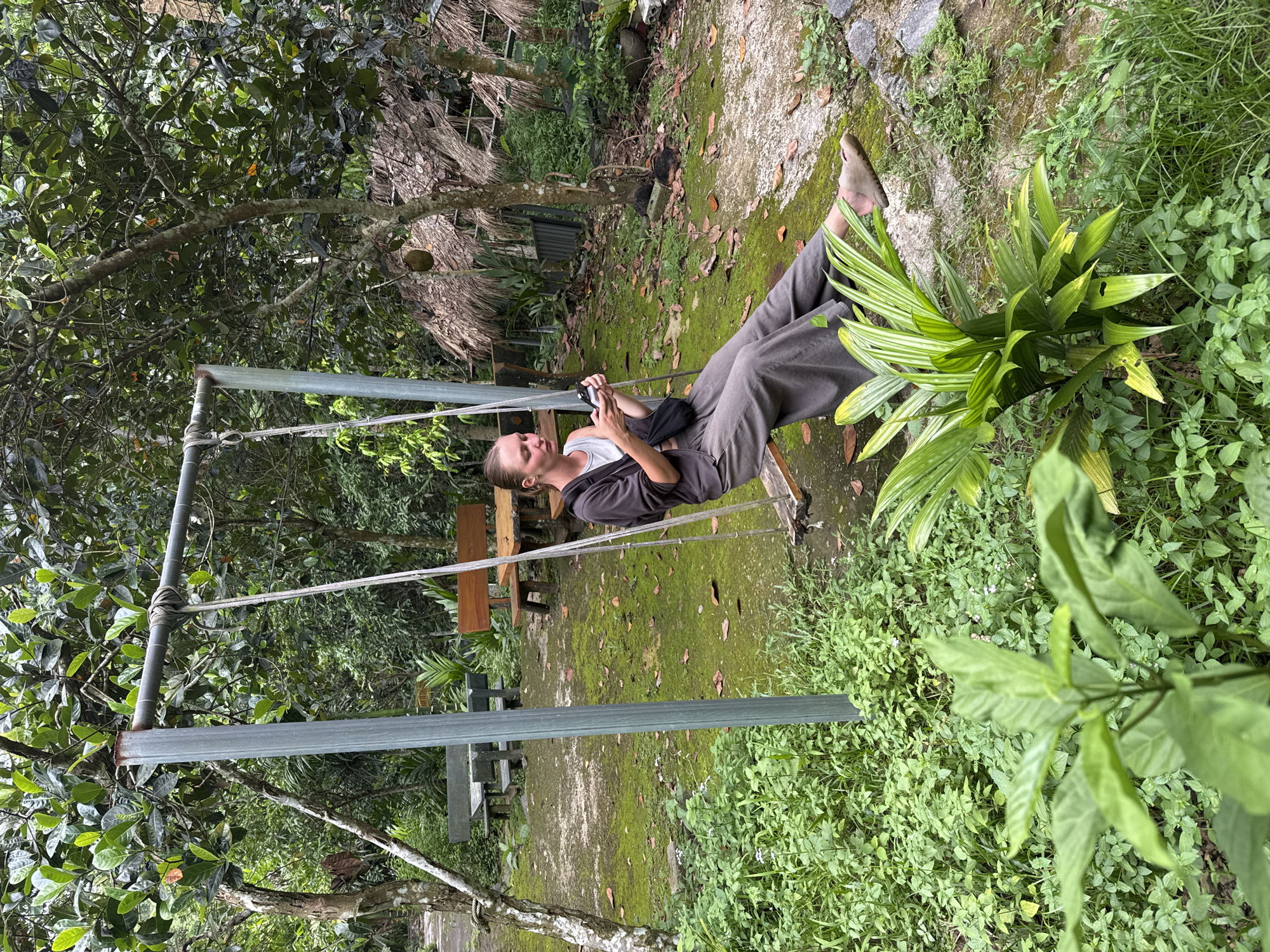 A young woman sits on a wooden swing in an outdoor setting with lush green vegetation. She is wearing a gray hooded jacket over a white tank top and loose gray pants. Her legs are extended, and she is barefoot. The swing is suspended by ropes from a metal frame. In the background, there are trees, thatched-roof structures, and park benches. The ground is covered in moss and fallen leaves. The location appears to be a park or a rural garden area within Cẩm Thạch Commune, Vietnam.