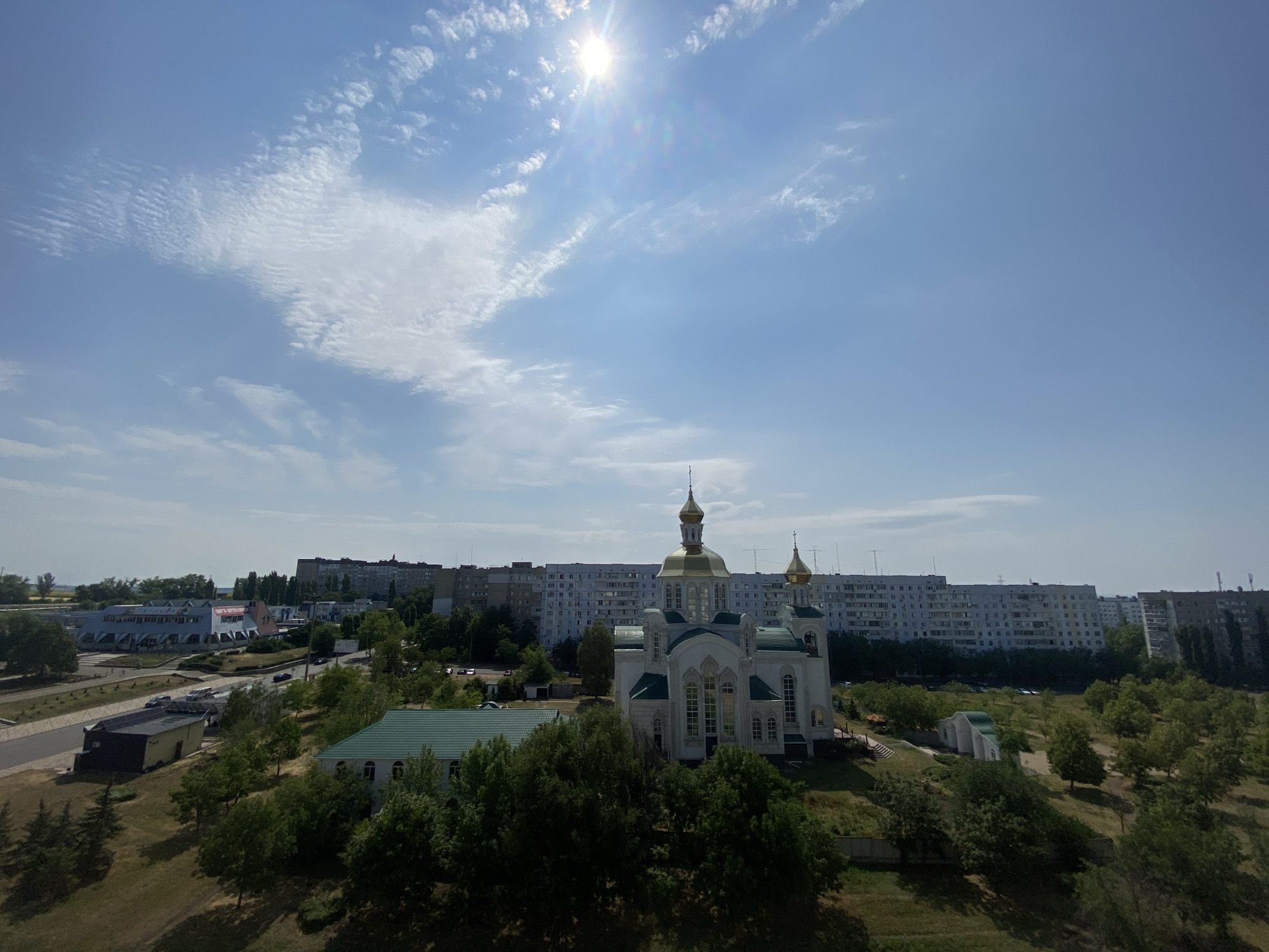 The image captures a bright, sunny day in Pivdennoukrainsk, Ukraine, with a clear blue sky and scattered white clouds. The sun is high in the sky, casting a bright glare. In the foreground, there is a white church with green roofing and golden domes, situated on a grassy hill surrounded by lush green trees. Behind the church, a row of tall, light-colored apartment buildings stretches across the horizon. To the left of the church, an industrial-looking building with a patterned facade is visible, as well as some smaller, darker buildings and parked cars. The overall scene depicts a residential area with a prominent religious building, common in many Ukrainian cities.