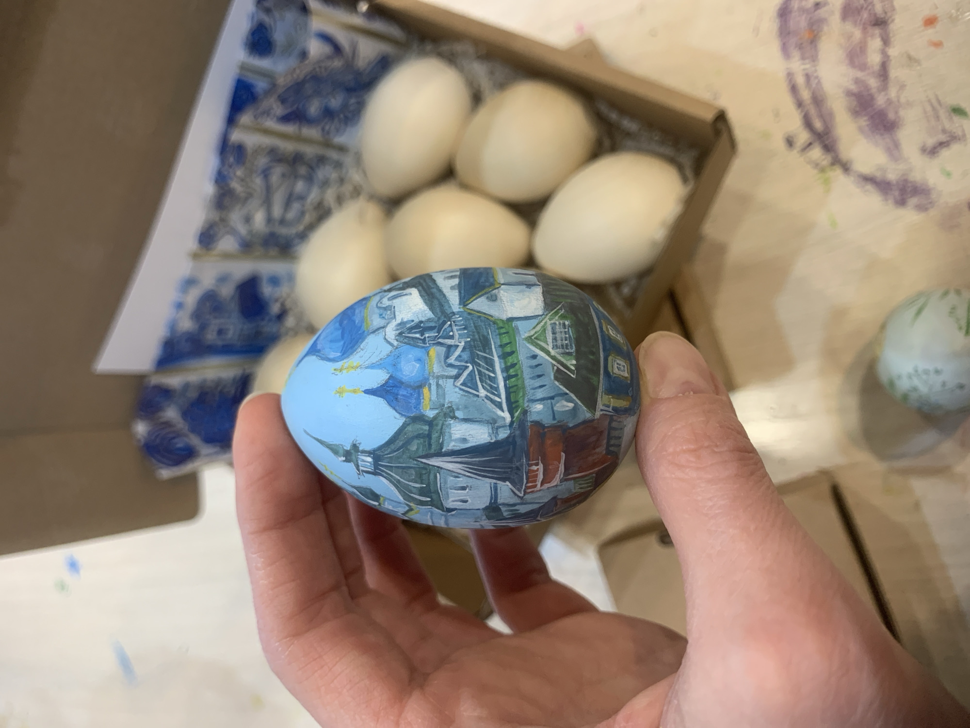A hand holds a painted egg. The egg is decorated with an image of architecture in blue, grey, red and white. Behind the egg is a cardboard box containing six plain white eggs. The box interior is lined with blue and white patterned paper. To the right is a partially visible painted egg. The background is a wooden surface with smudges of paint.