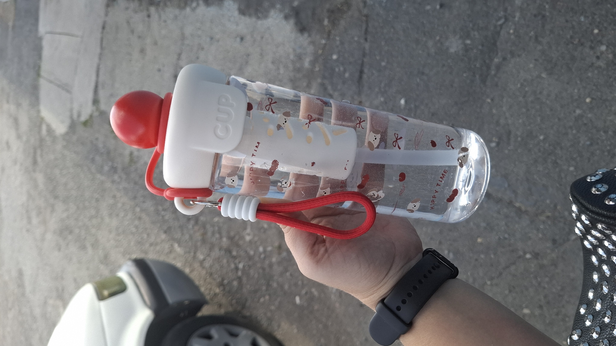 A person's left hand, adorned with a black smartwatch, holds a clear plastic water bottle. The bottle is decorated with images of cartoon dogs, red cherries, and bows, and the words "HAPPY TIME" are visible. A white straw extends from the top of the bottle, which is capped with a white lid and a red spherical knob. A red cord with white accents is attached to the lid. In the background, the front of a white car is partially visible, and the ground is paved with asphalt. The scene is likely outdoors, possibly in a residential area or parking lot. The lighting suggests it is daytime.