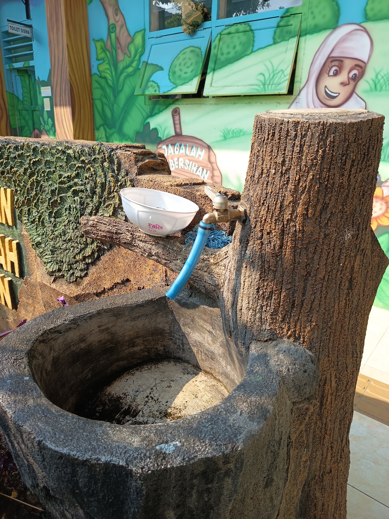 This image depicts an outdoor sink area, possibly located in Widoro, Indonesia, situated near a wall decorated with colorful murals. The murals feature cartoonish depictions of nature, including trees, leaves, and a smiling girl in a headscarf.

The sink itself is a rustic, stone-like structure designed to resemble a tree stump. A faucet is mounted on a faux wood surface, with a blue hose attached, directing water into the stone basin. A white plastic bowl, with the word "FARA" inscribed on it, rests on the edge of the sink structure, near the faucet. The basin of the sink appears to contain some water and debris.

To the left of the sink, a textured, green wall element with prominent yellow letters spelling "JAGALAH BERSIHAN" (meaning "Keep Clean" in Indonesian) is visible. Above this, a sign reads "TOILET SISWA" ("Student Toilet"). The overall setting suggests an educational or public space with an emphasis on hygiene and environmental awareness. The lighting indicates it is daytime.